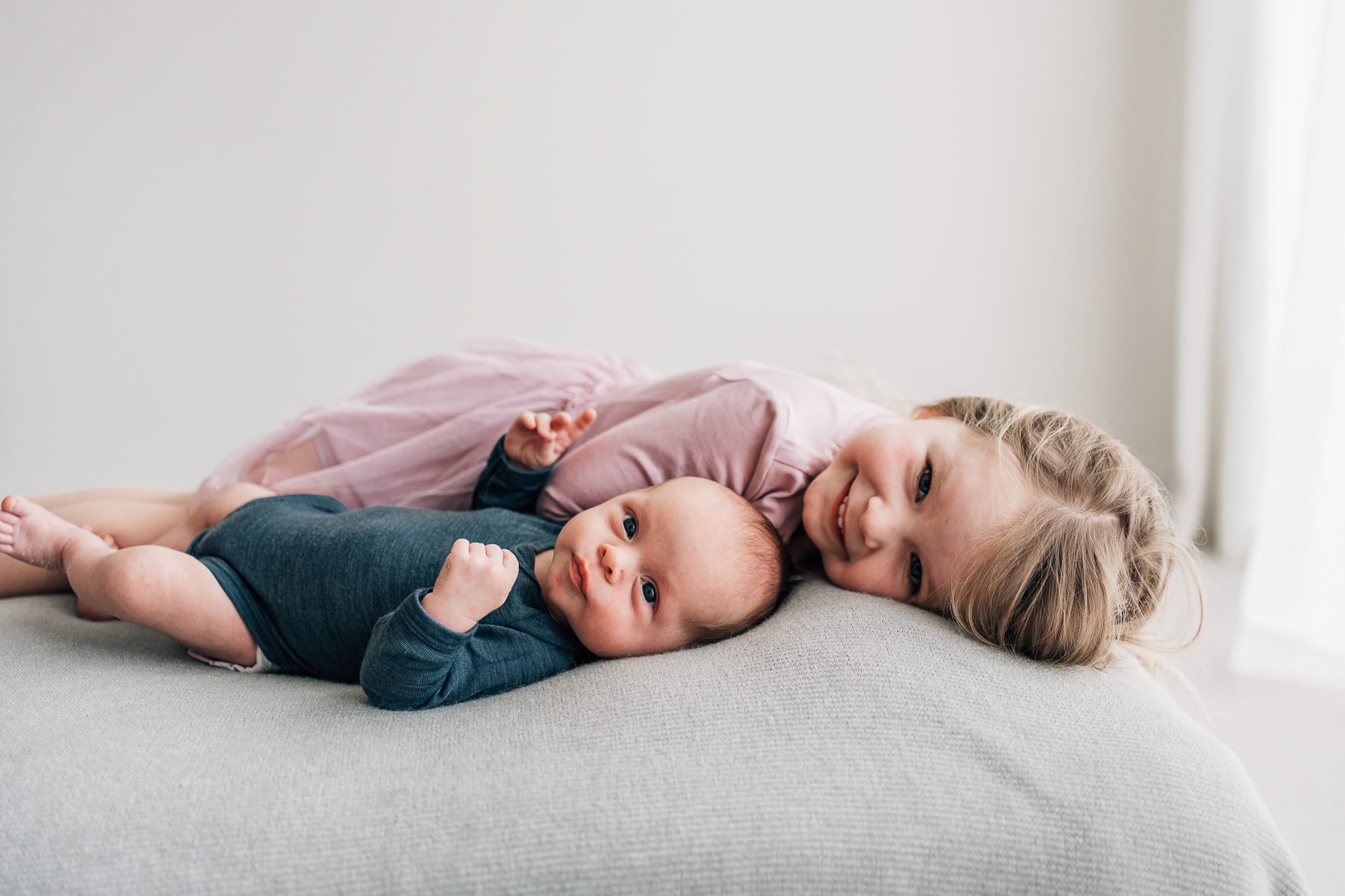 A smiling big sister lying down beside her newborn sibling.
