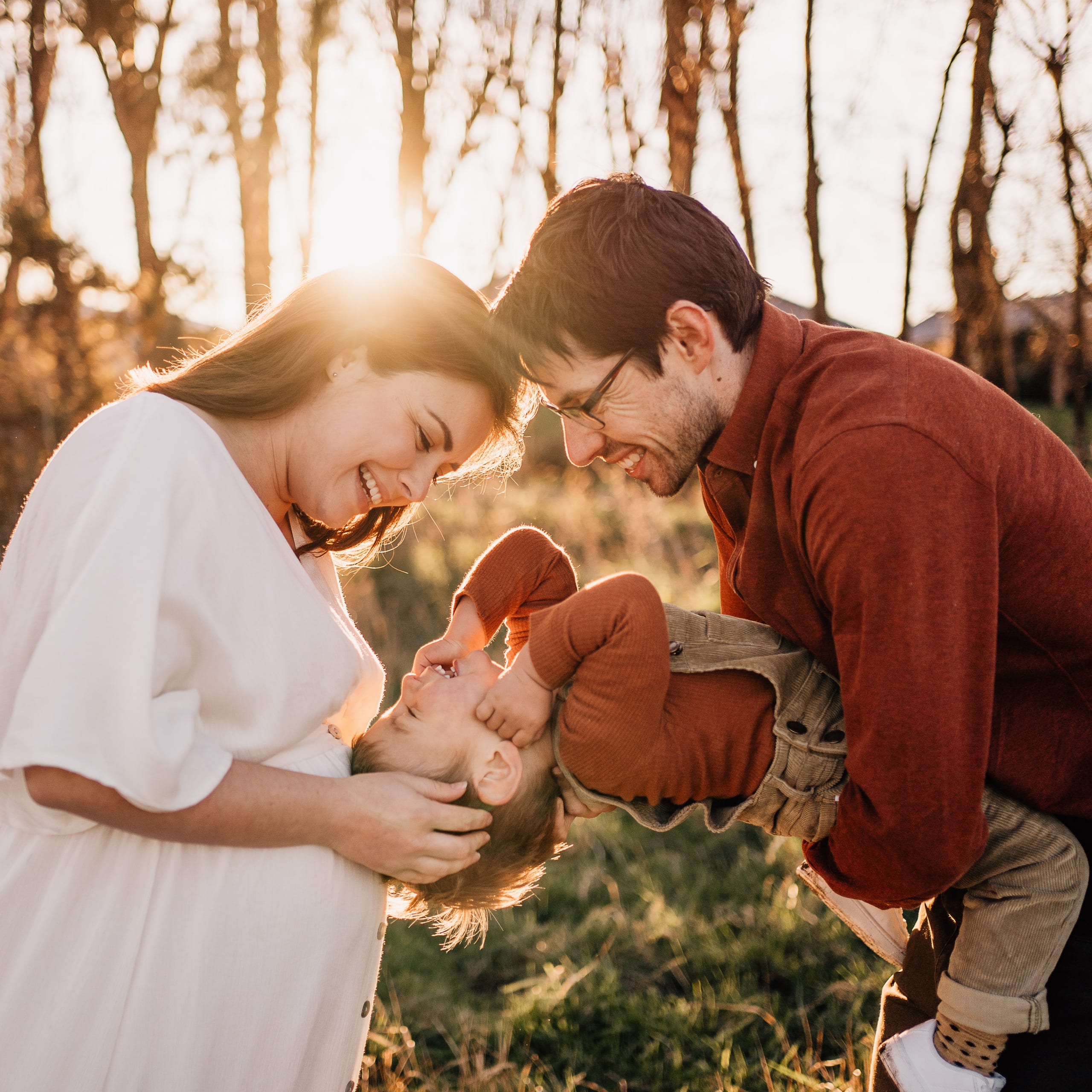 A pregnant mother and father sharing a laugh with their toddler in a golden field at sunset.