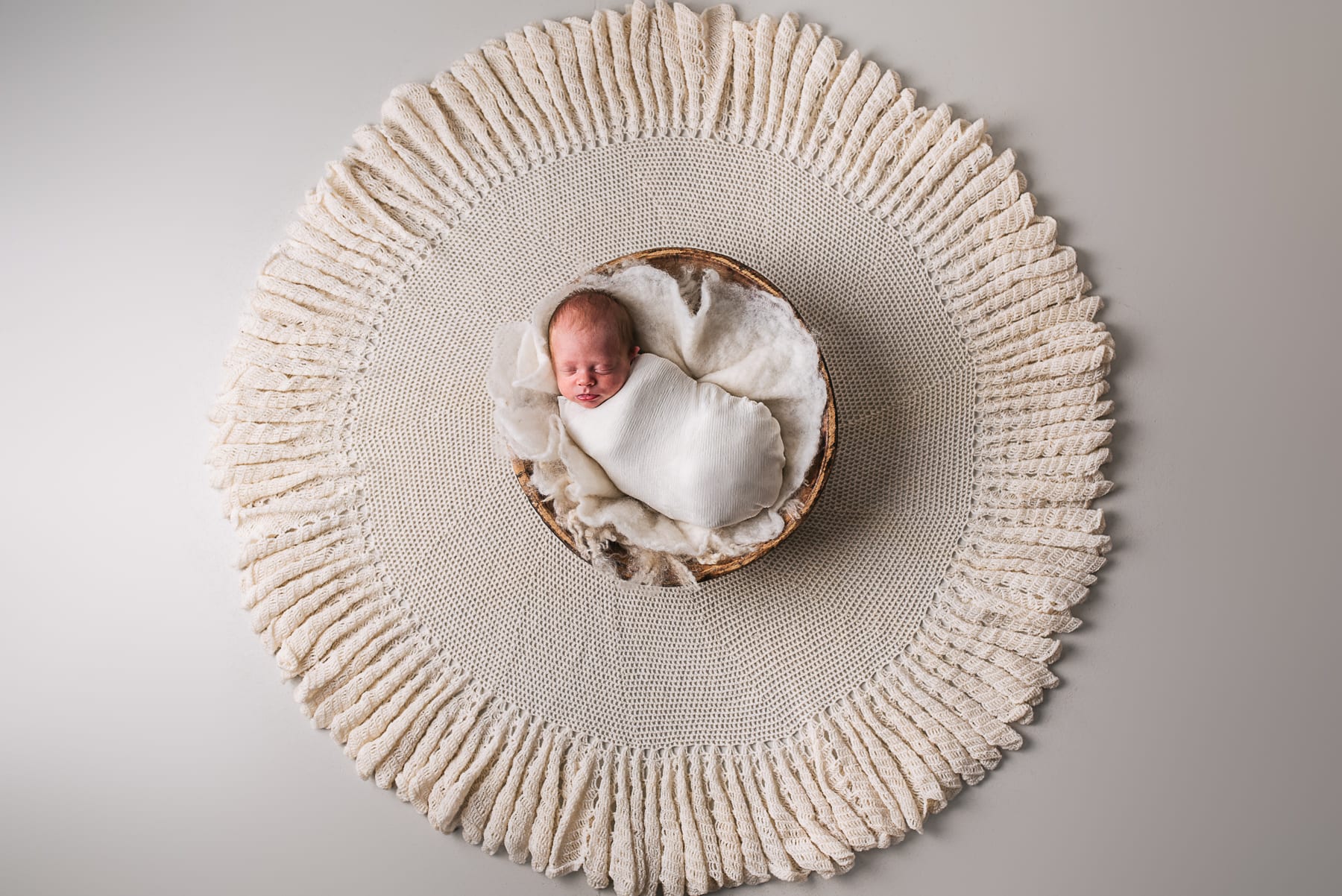 Overhead view of a swaddled newborn asleep in a wooden bowl on a soft cream knit.