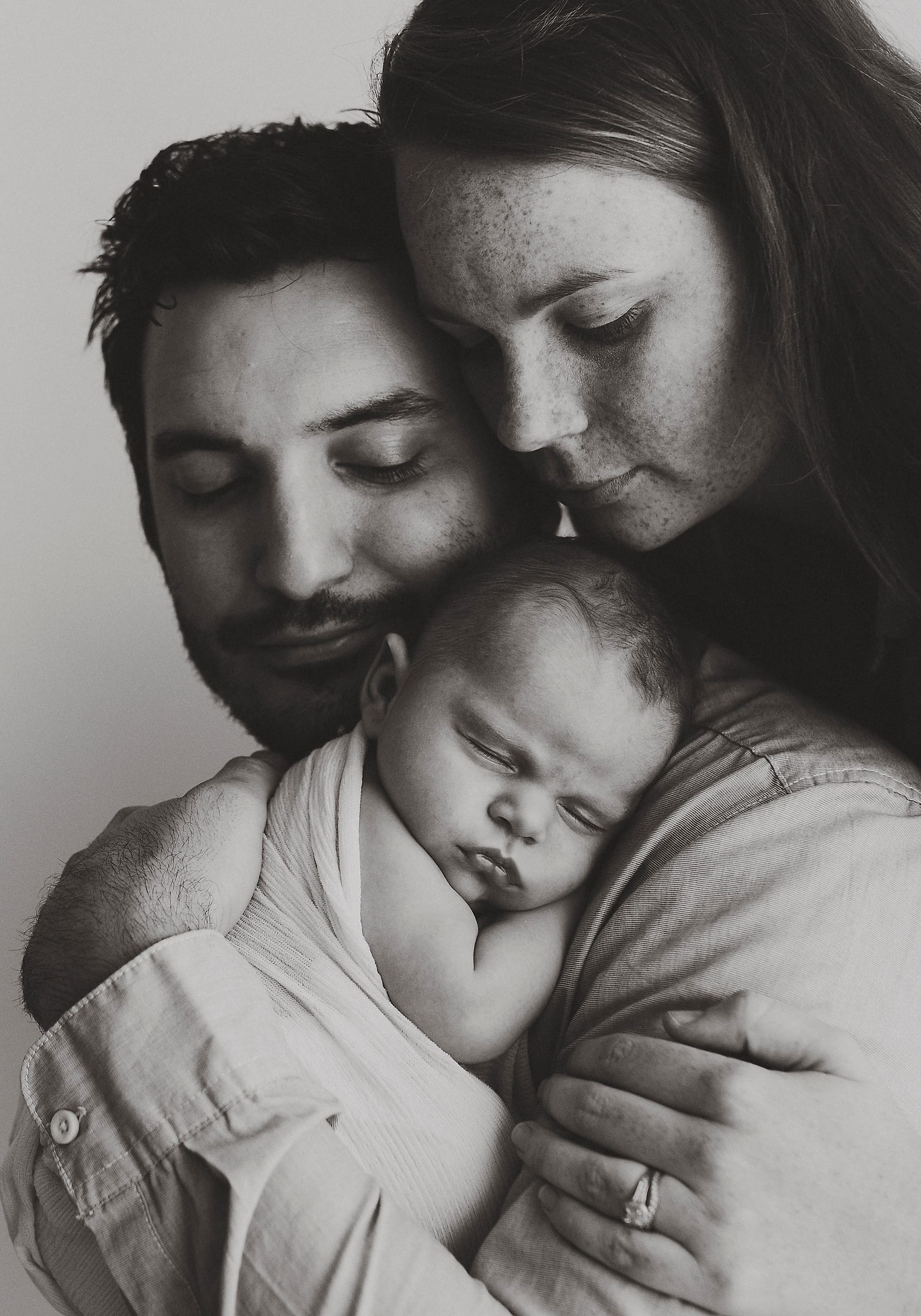 Black and white portrait of two parents cradling their sleeping newborn between them.