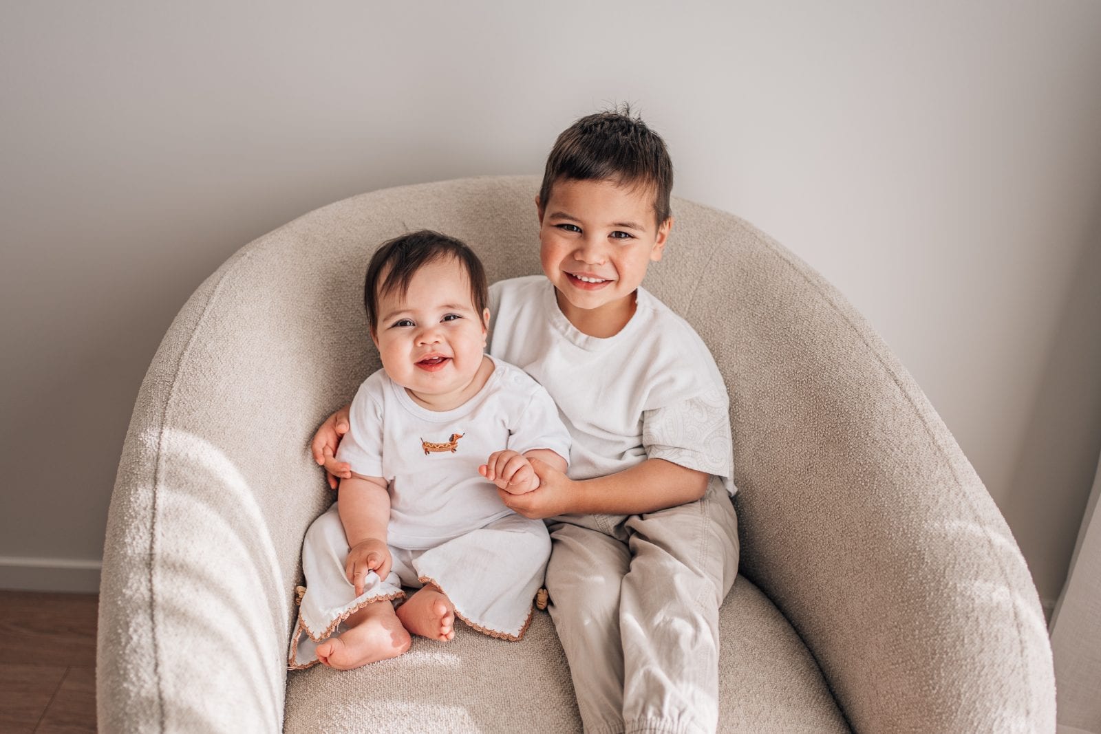 Smiling baby and big brother sitting together on a cream armchair in a bright natural-light studio.