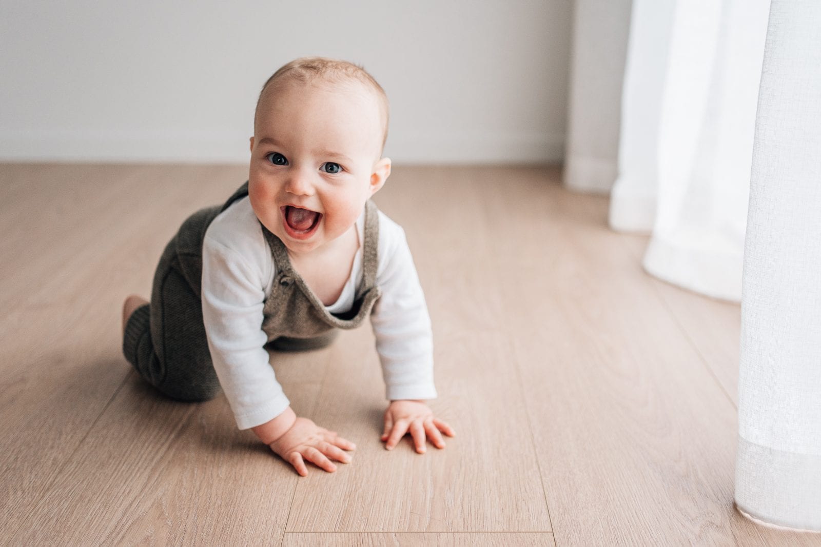 Grinning baby boy in knitted overalls crawling across the natural-light studio floor.