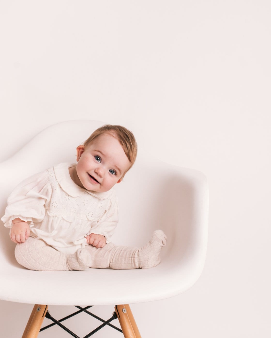 A baby in a cream outfit sits smiling on a white designer chair during a Little Portrait Club studio session.