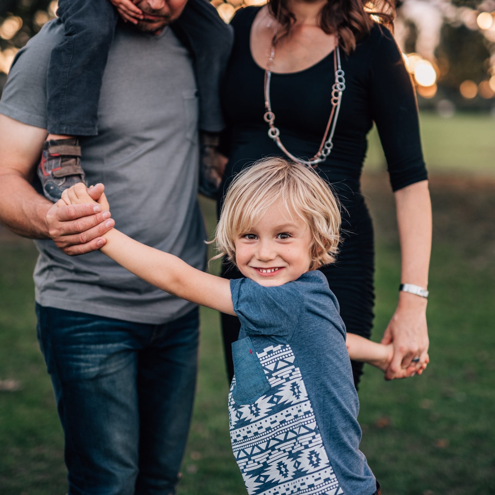 A grinning blonde boy holds his parents' hands as the family walks together across a grassy park at dusk.