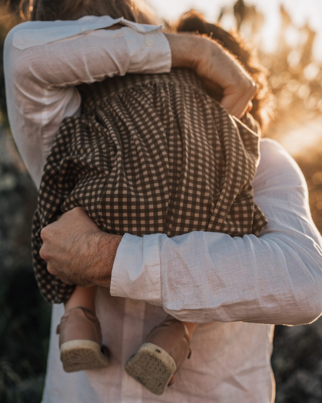 A father in a white linen shirt cradles his toddler daughter in a gingham dress, backlit by warm golden sunset light.