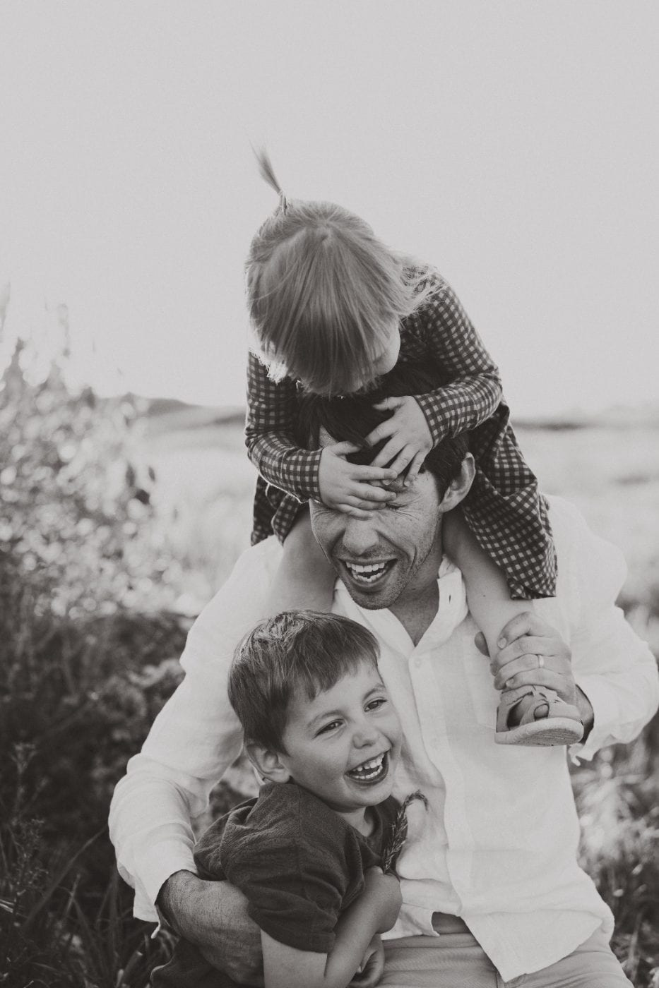 Black and white candid of a laughing dad with his young daughter on his shoulders and son in his lap among tall grass.