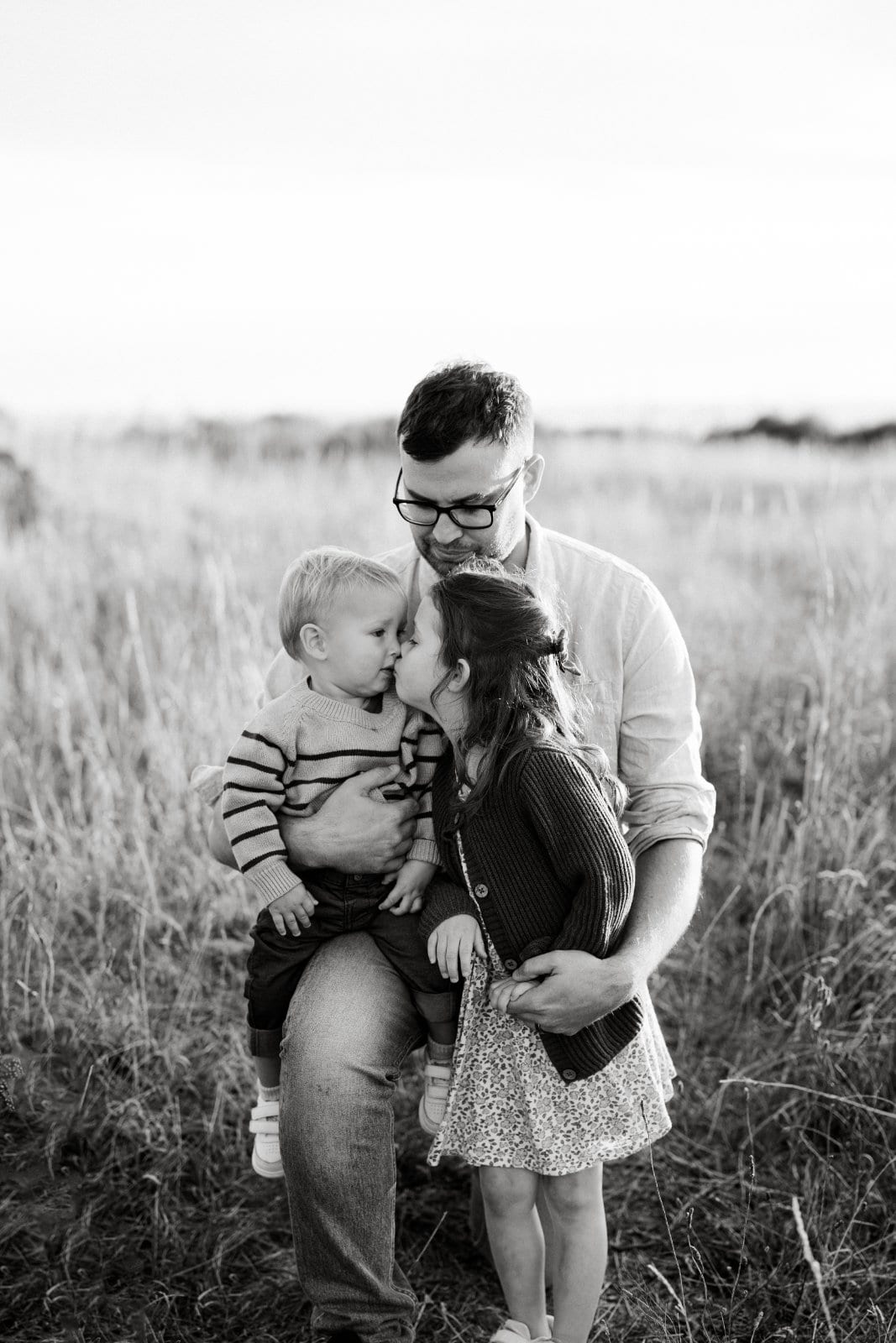 Black and white portrait of a father holding his son and daughter as they kiss cheeks in a grassy field.