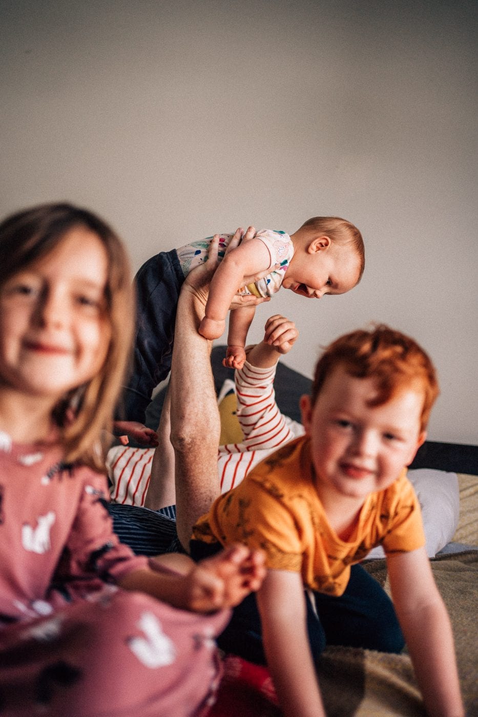A father lifts his laughing baby in the air while the two older siblings sit smiling on the bed.