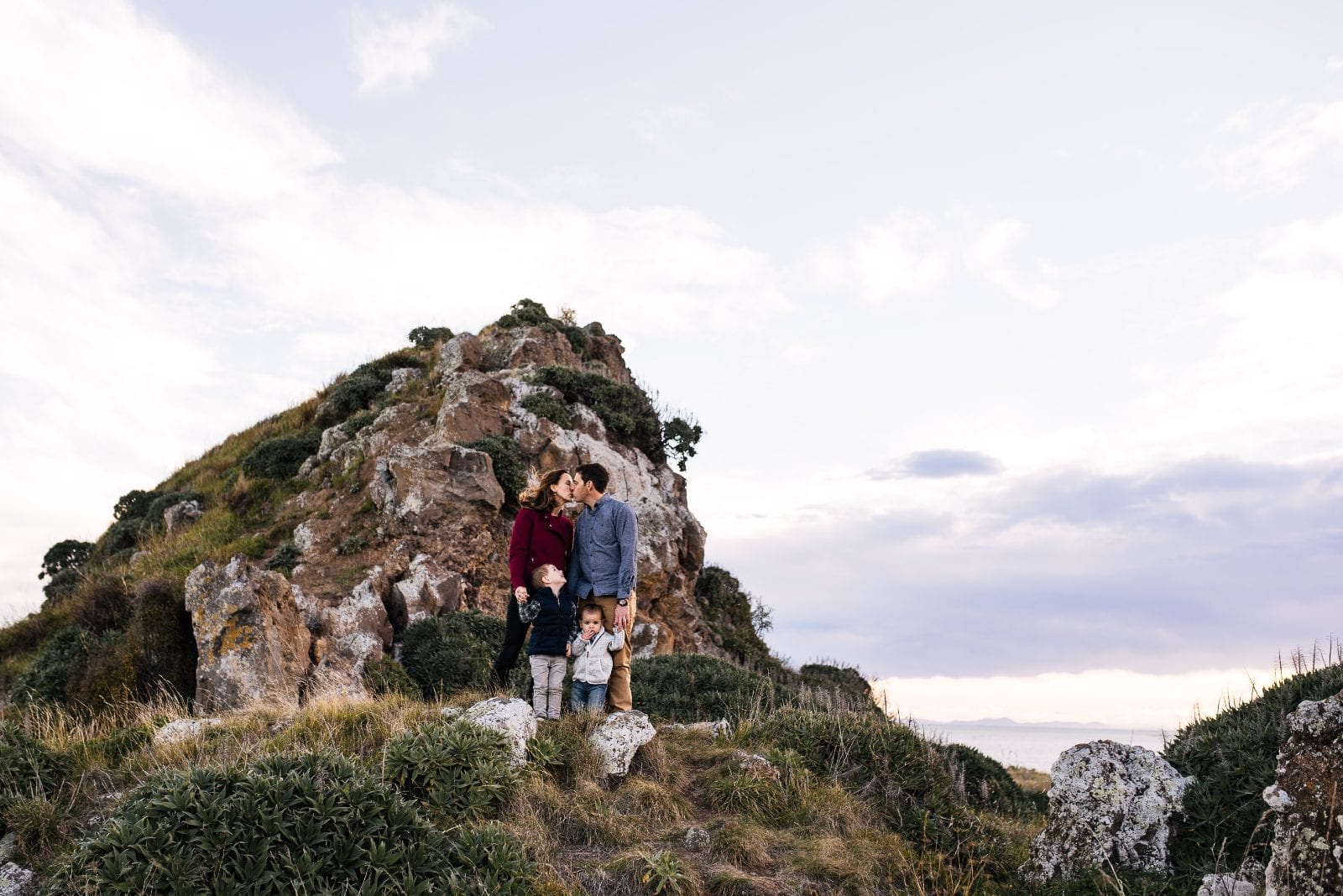 Parents kiss while standing with their two young sons on a rocky coastal headland with the sea beyond.