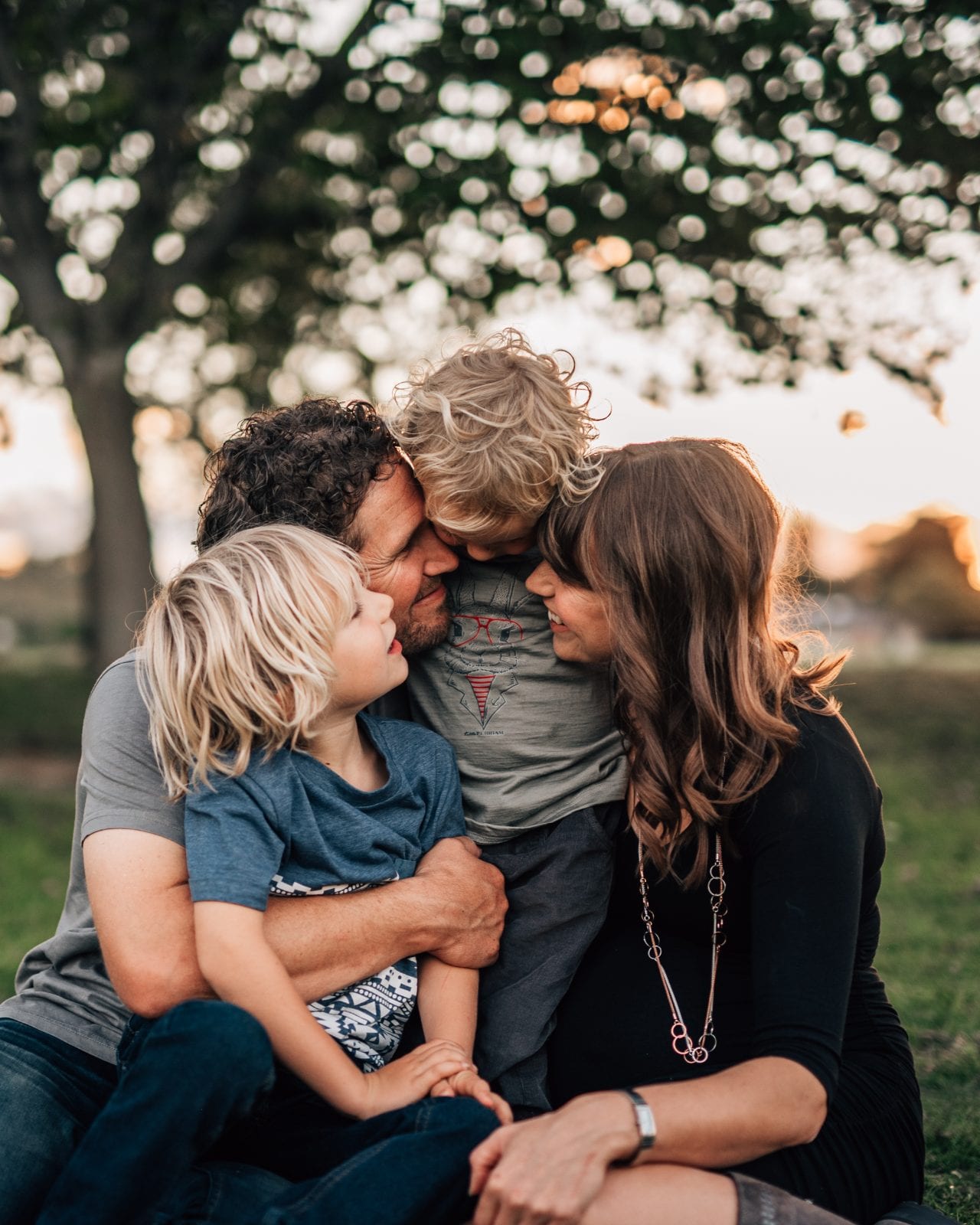 A father and mother cuddle their two young sons together in a park at sunset with soft glowing bokeh behind.