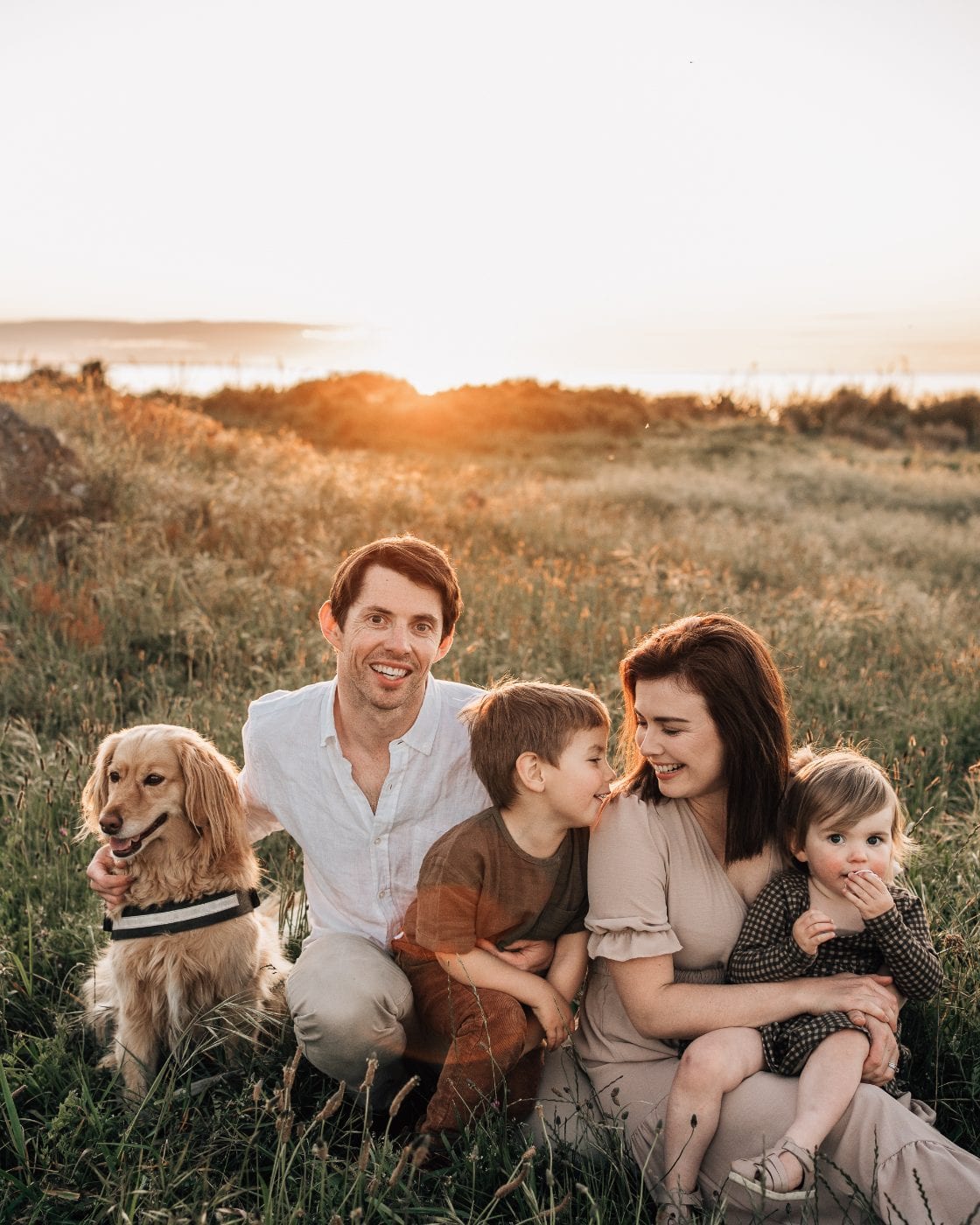 A family of four with their golden retriever sit smiling together in a coastal meadow as the sun sets behind them.