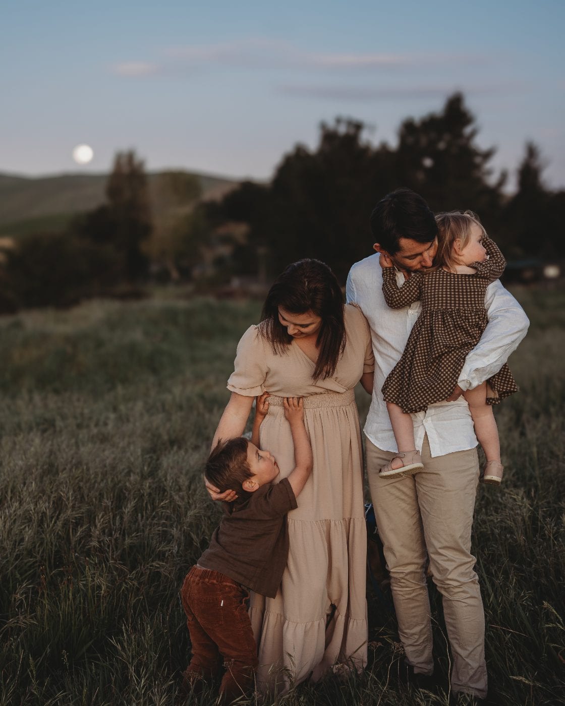Parents and their two young children embrace in a grassy field at dusk while the father kisses his daughter and the moon rises.