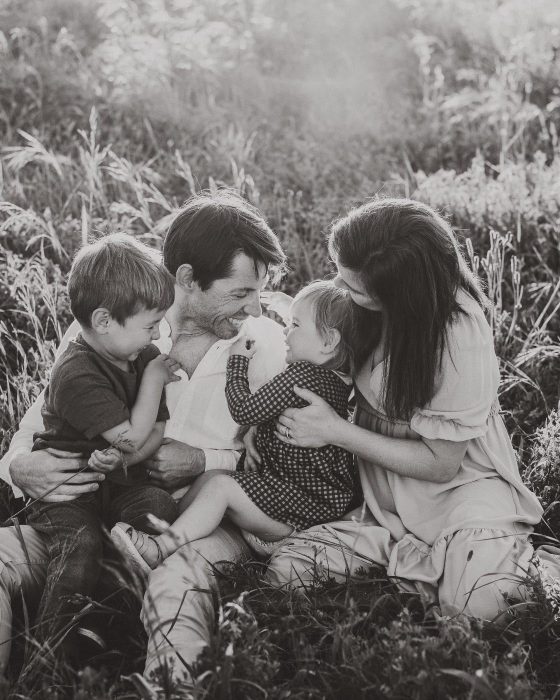 Black and white image of a family of four laughing and playing together while sitting in a sunlit meadow of tall grass.