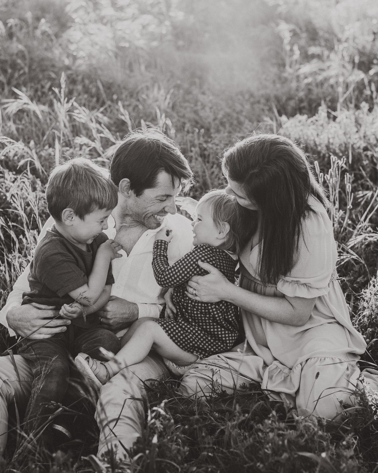 Black-and-white portrait of parents and two children laughing together as they sit among tall meadow grass.