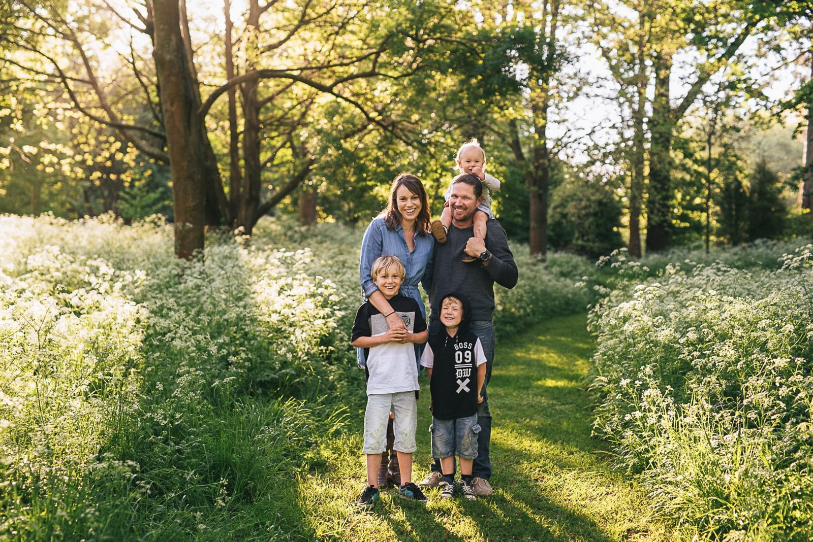 A family of five smiles together on a grassy path surrounded by white cow parsley in a sunlit woodland.