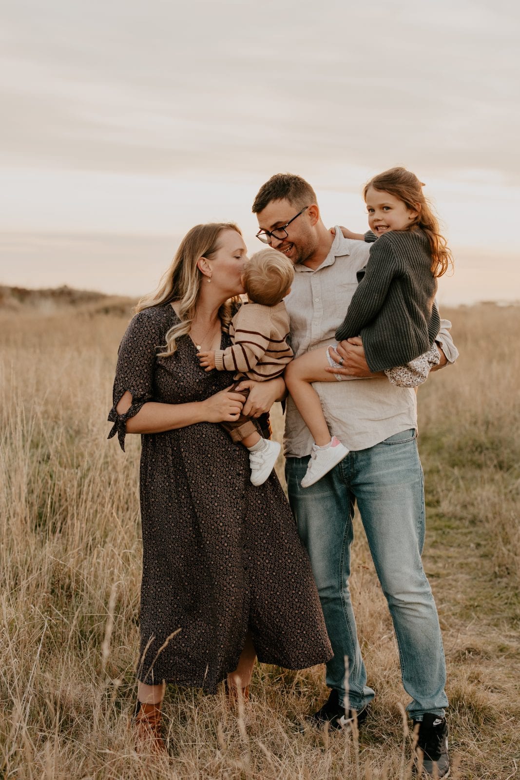 Parents share a kiss with their toddler while holding their two young children in a golden field at sunset.