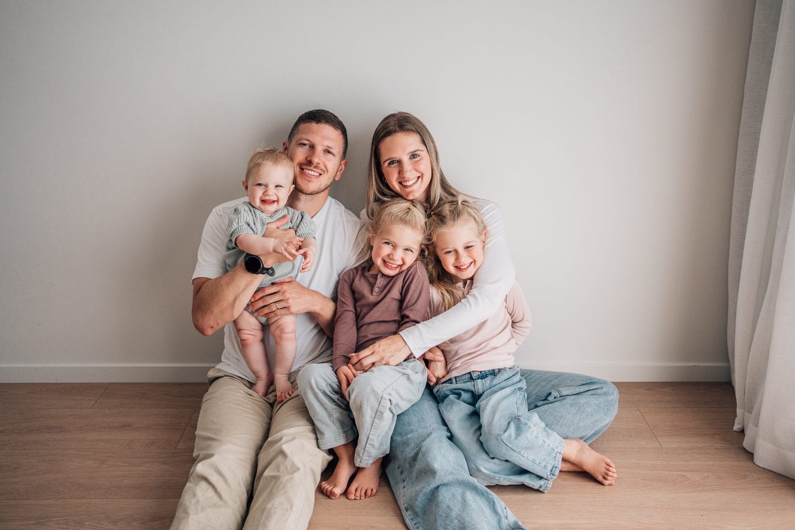 Parents with their baby and two young daughters smiling together in a natural-light family studio portrait.