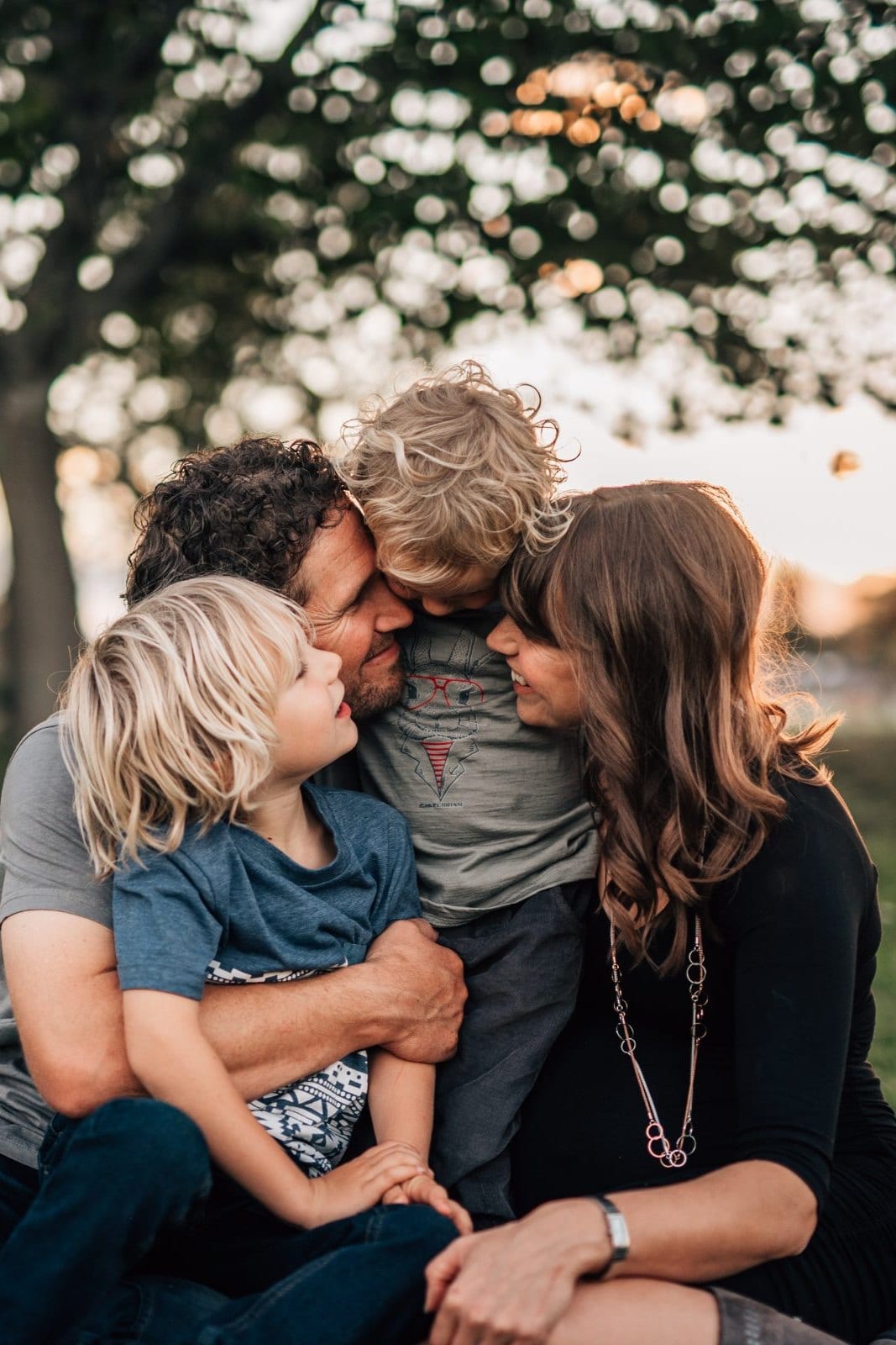 Parents and two young sons share a close, laughing embrace beneath a tree at sunset in a leafy park.
