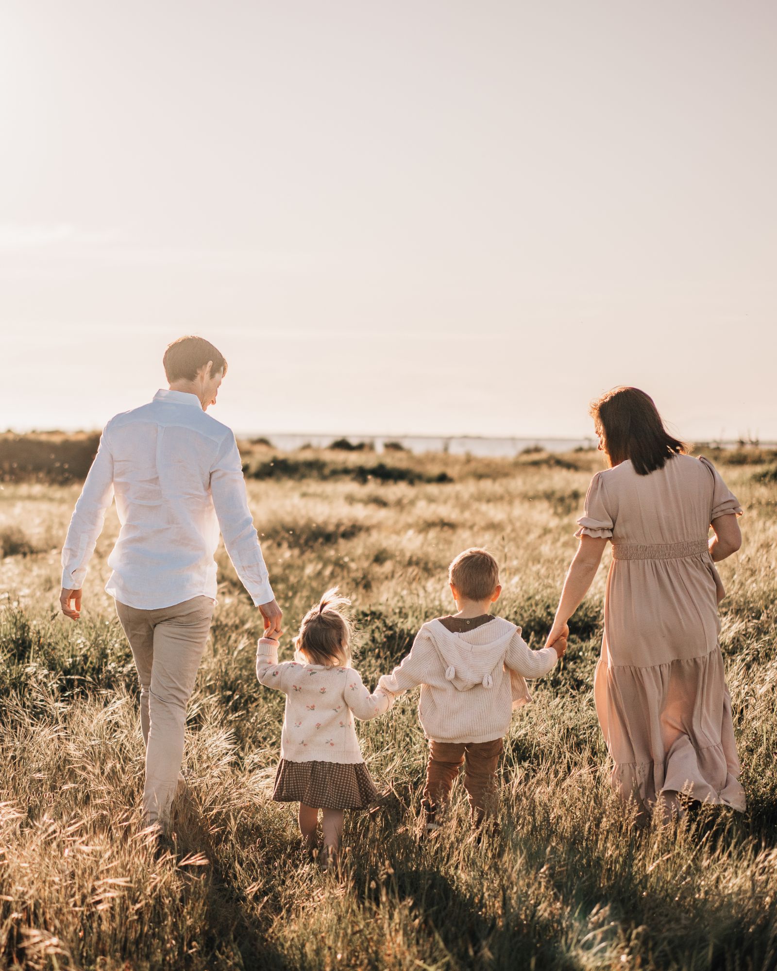 Family of four walking hand in hand through golden grass at sunset, dressed in warm earthy tones