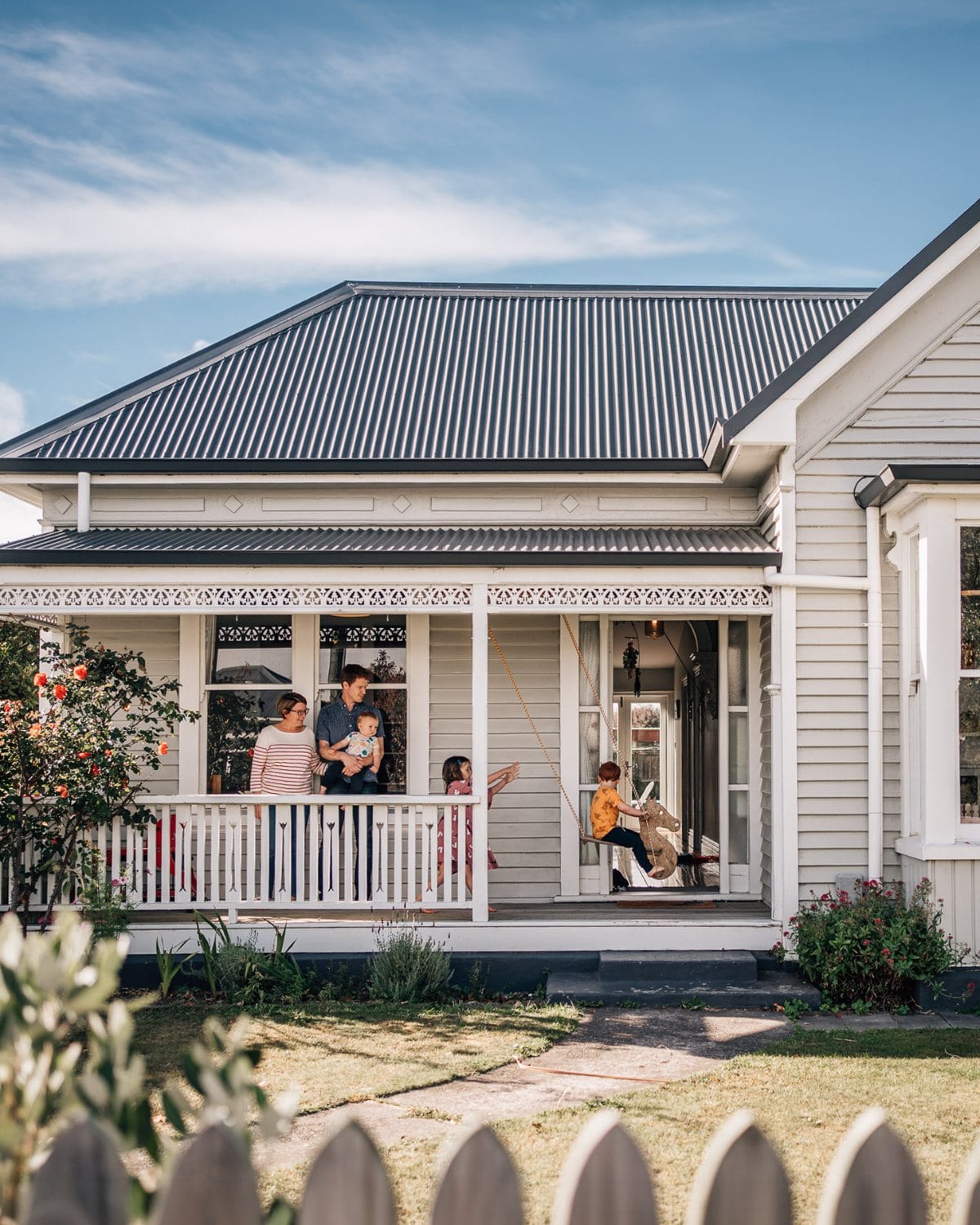 A family gathers on the veranda of a white weatherboard villa while two children play on a rope swing in the sunny garden.