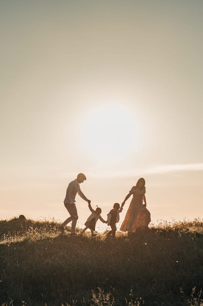 Silhouetted parents and two children hold hands and play together on a grassy hilltop against a glowing sunset sky.