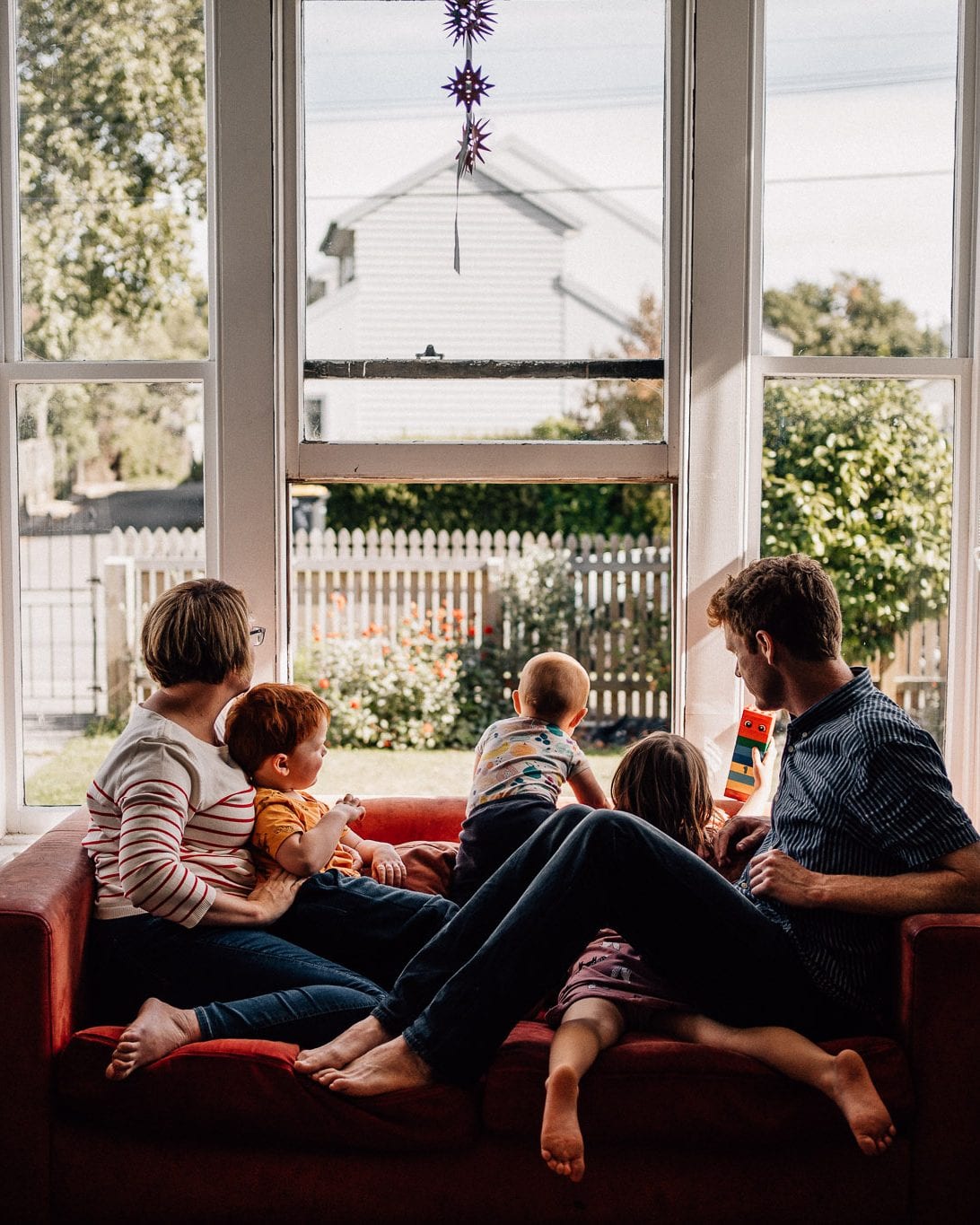 A family of six relaxes together on a red sofa by a sunny bay window looking out to the garden.