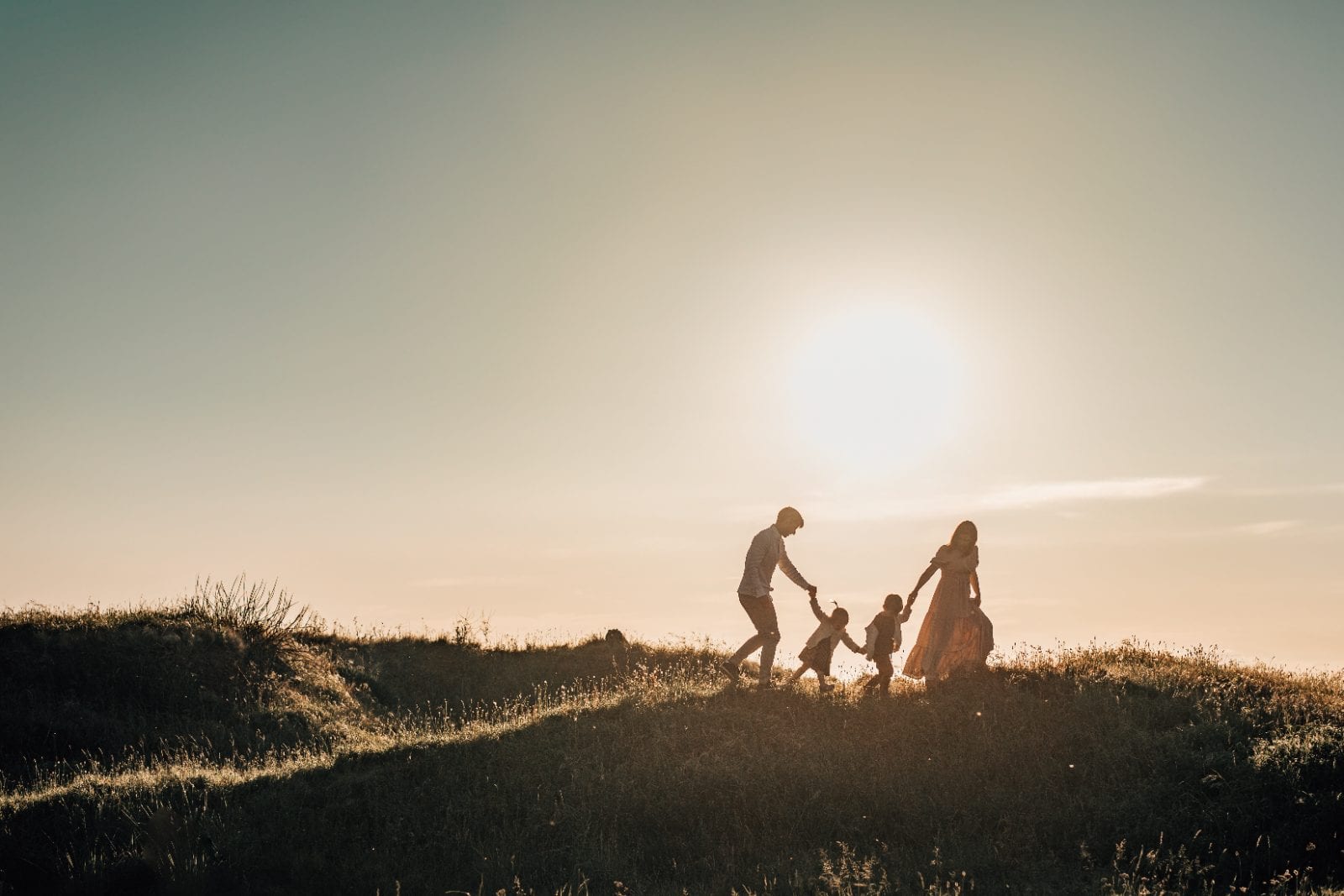 Silhouetted family joins hands to play ring-a-rosie on a grassy ridge beneath a radiant setting sun.