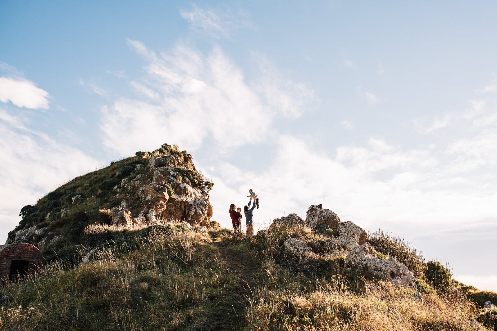 A family stands on a rocky grassy outcrop under a vast blue sky as a father lifts his child into the air.