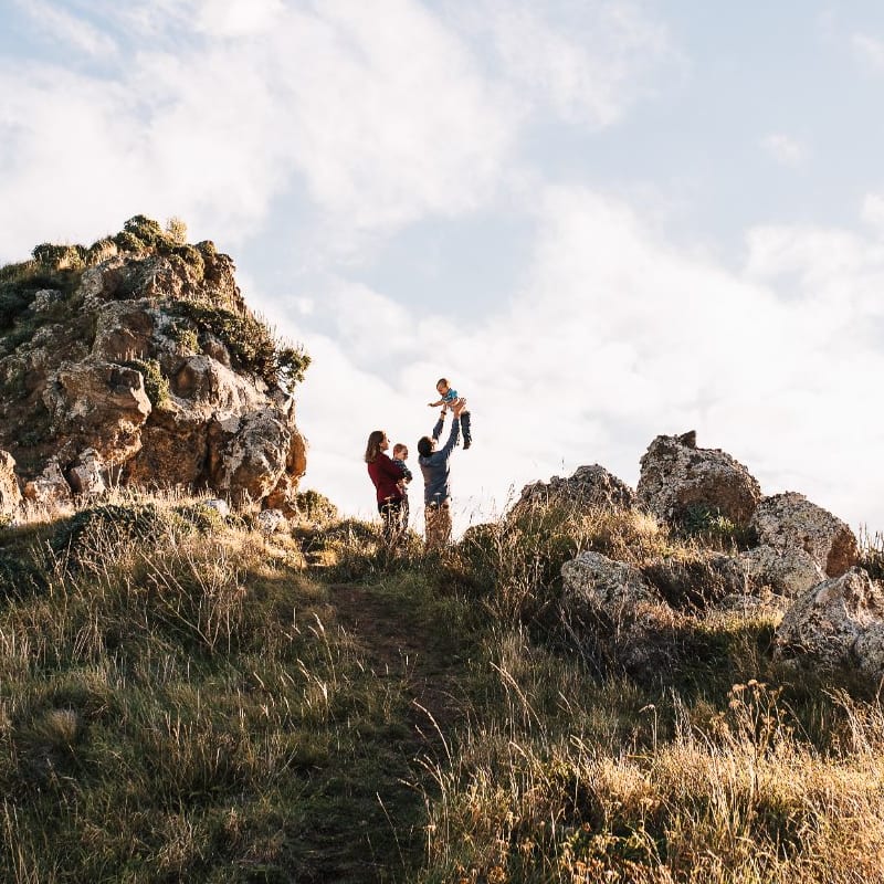 A family plays on a rugged grassy hilltop among rocks, lifting a small child up as the sun lowers.