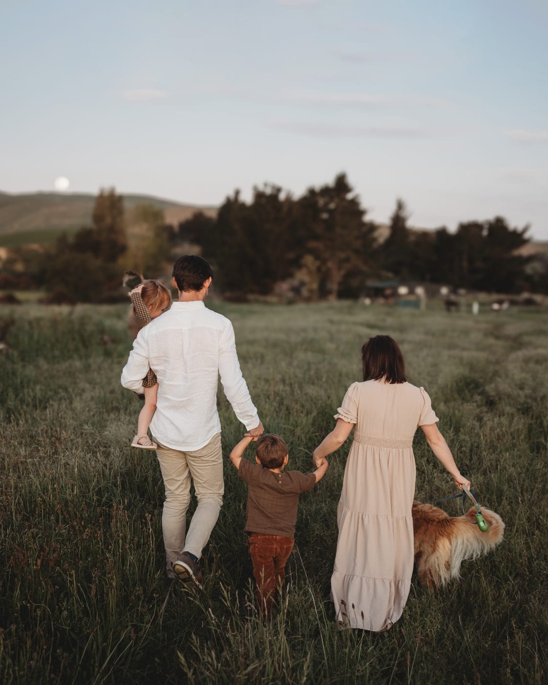 A family of four and their golden retriever walk away hand-in-hand through a grassy Canterbury field as the moon rises over the hills at dusk.
