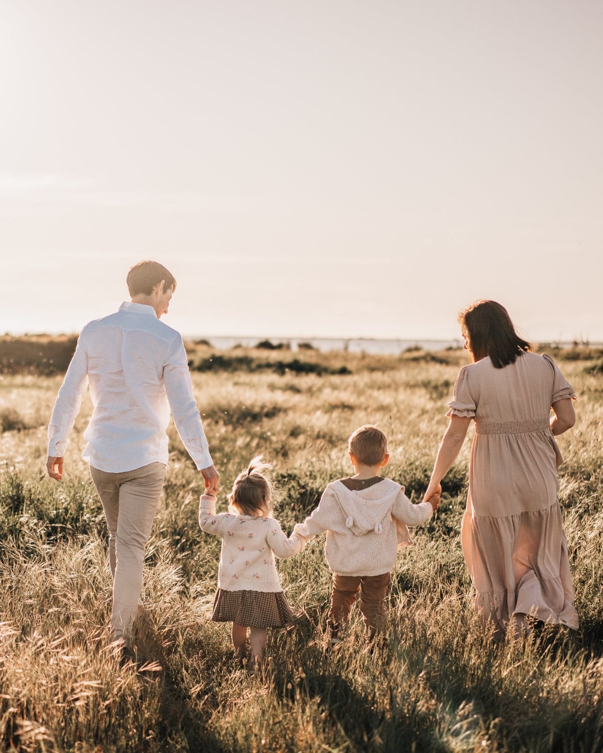 A mother and father walk away hand in hand with their two children through tall grass in warm evening light.