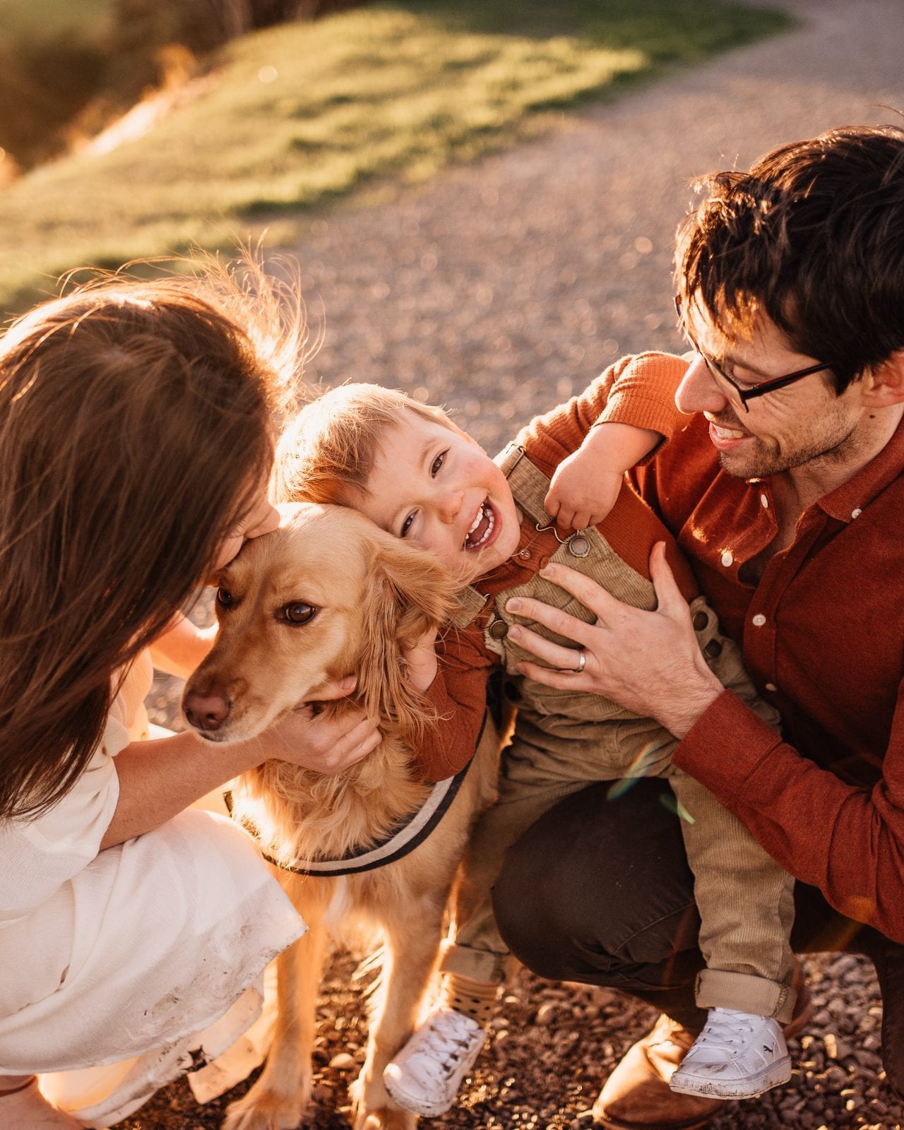 A toddler laughing between their parents while cuddling the family's golden dog at golden hour.