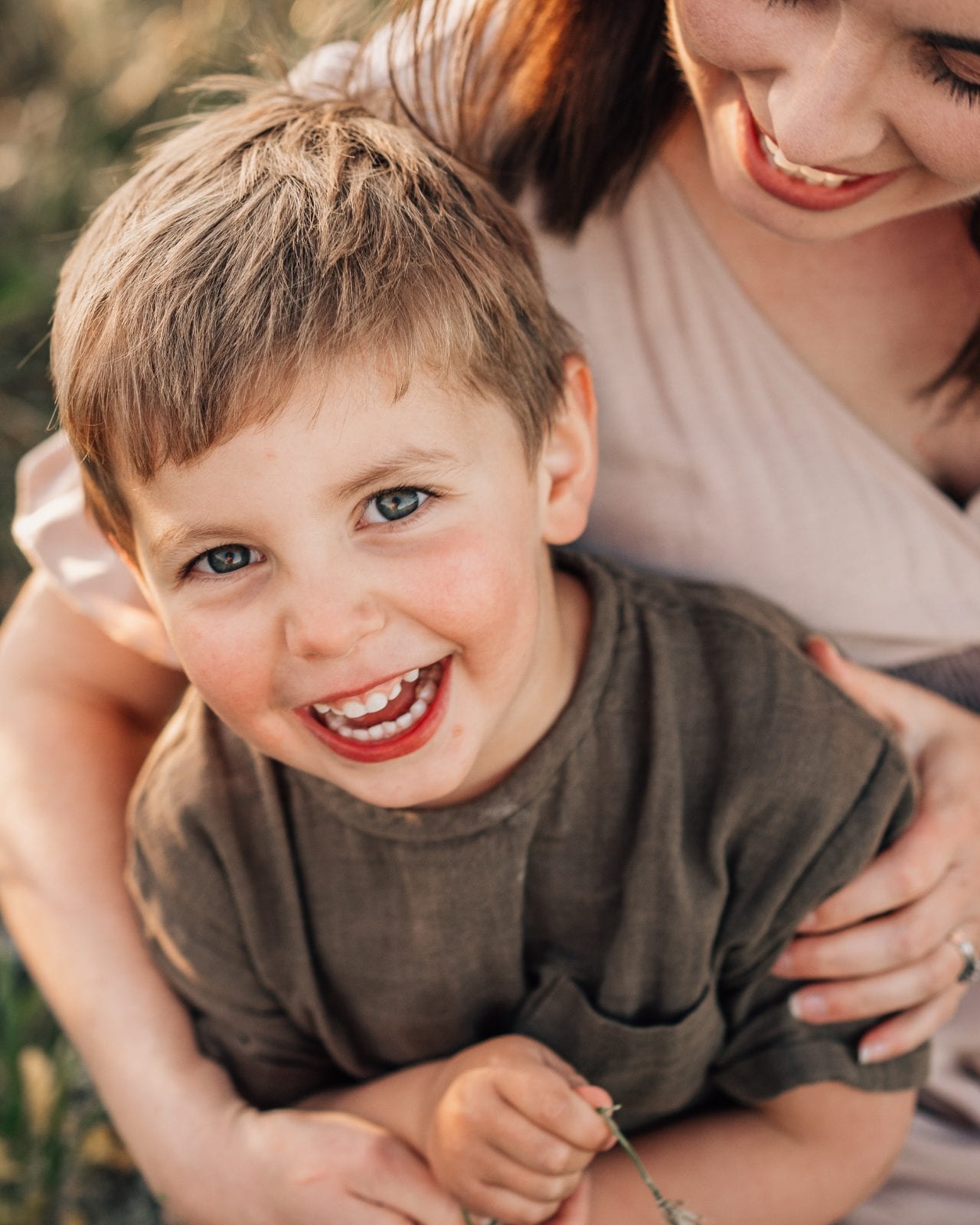 A bright-eyed young boy laughs joyfully while his mother holds him close in a sunlit grassy field.