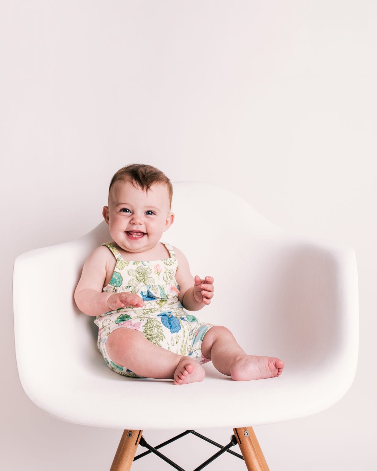 A laughing baby in a botanical print romper sits on a white designer chair.