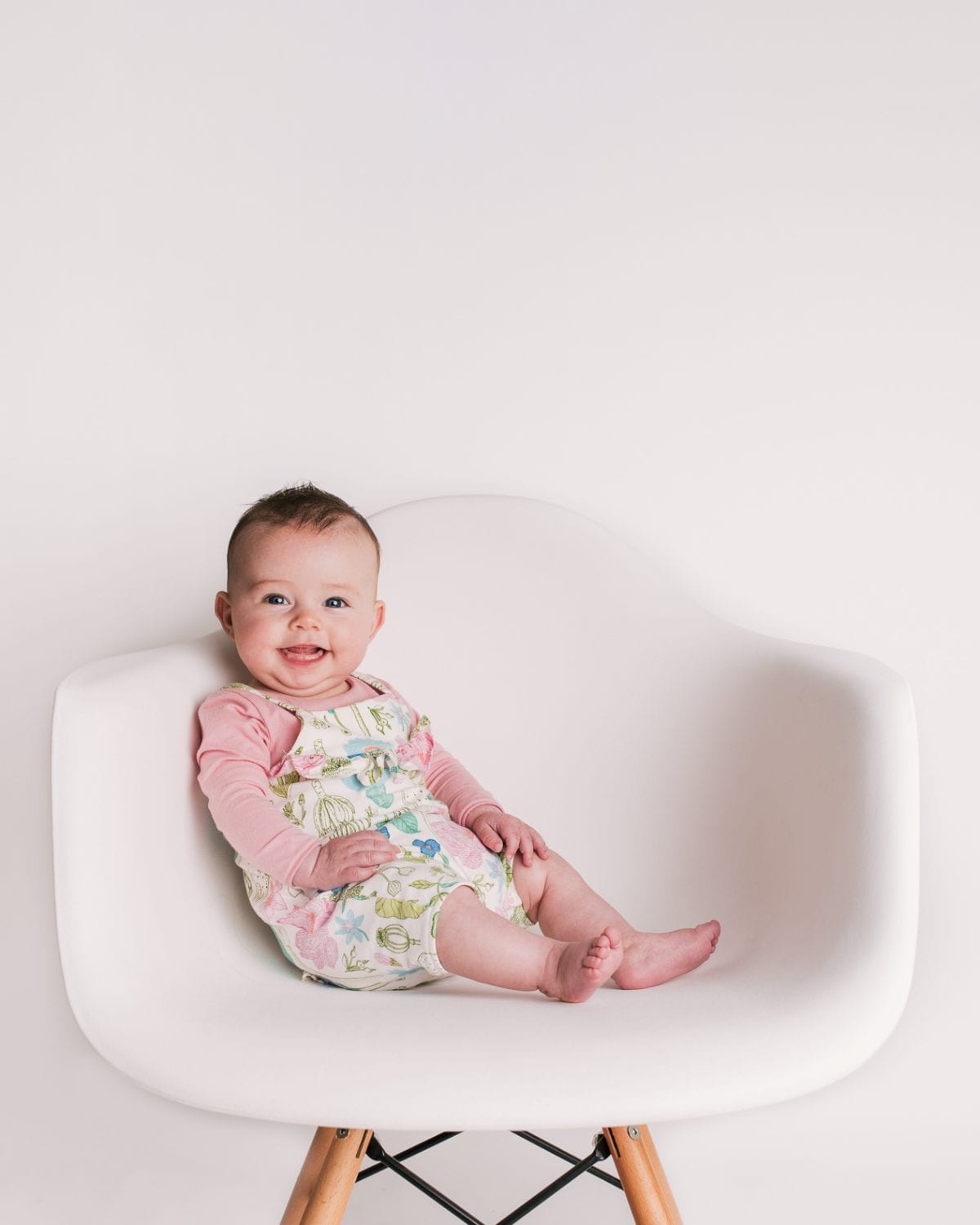 A smiling baby in a floral romper with pink sleeves sits on a white designer chair.