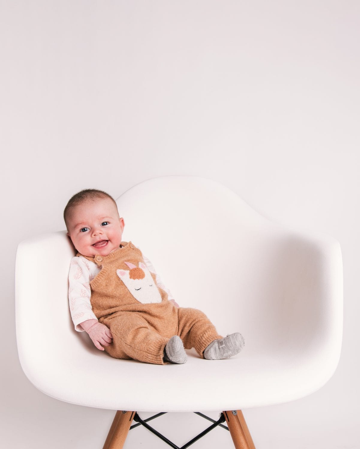 A smiling baby in mustard horse dungarees reclines on a white designer chair.