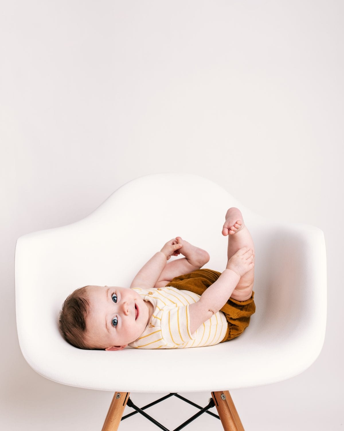 A baby in a striped top and mustard shorts lies back with feet up on a white chair.