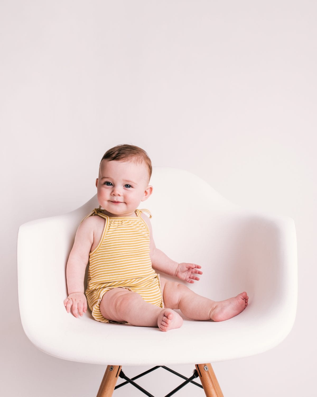 A baby in a yellow striped romper sits on a white designer chair.