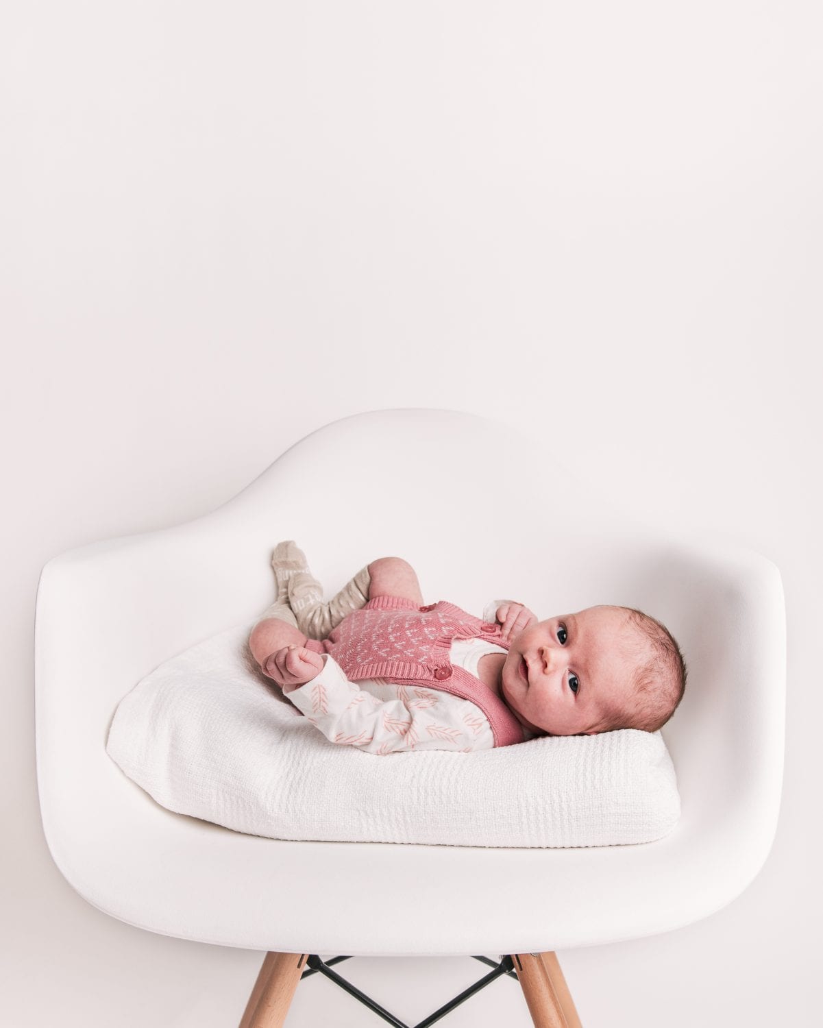 A newborn in a pink knit vest rests on a cushion on a white chair.