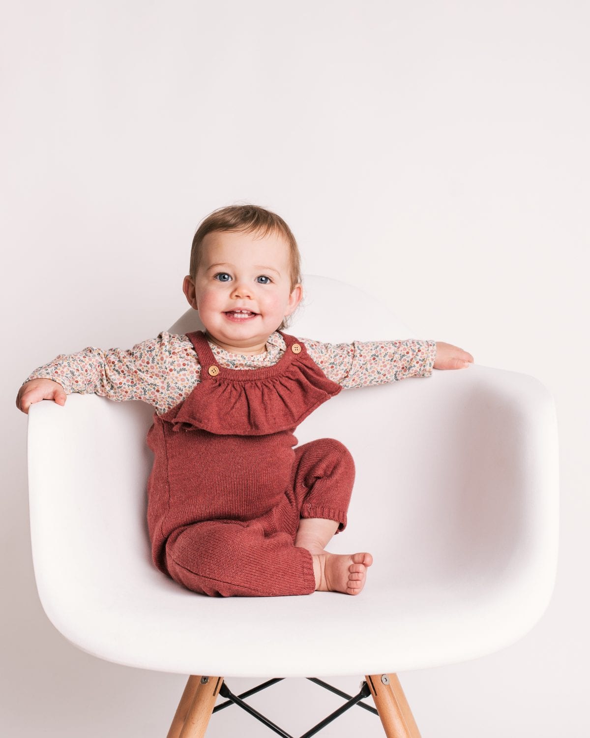 A grinning toddler in rust dungarees spreads their arms across a white designer chair.