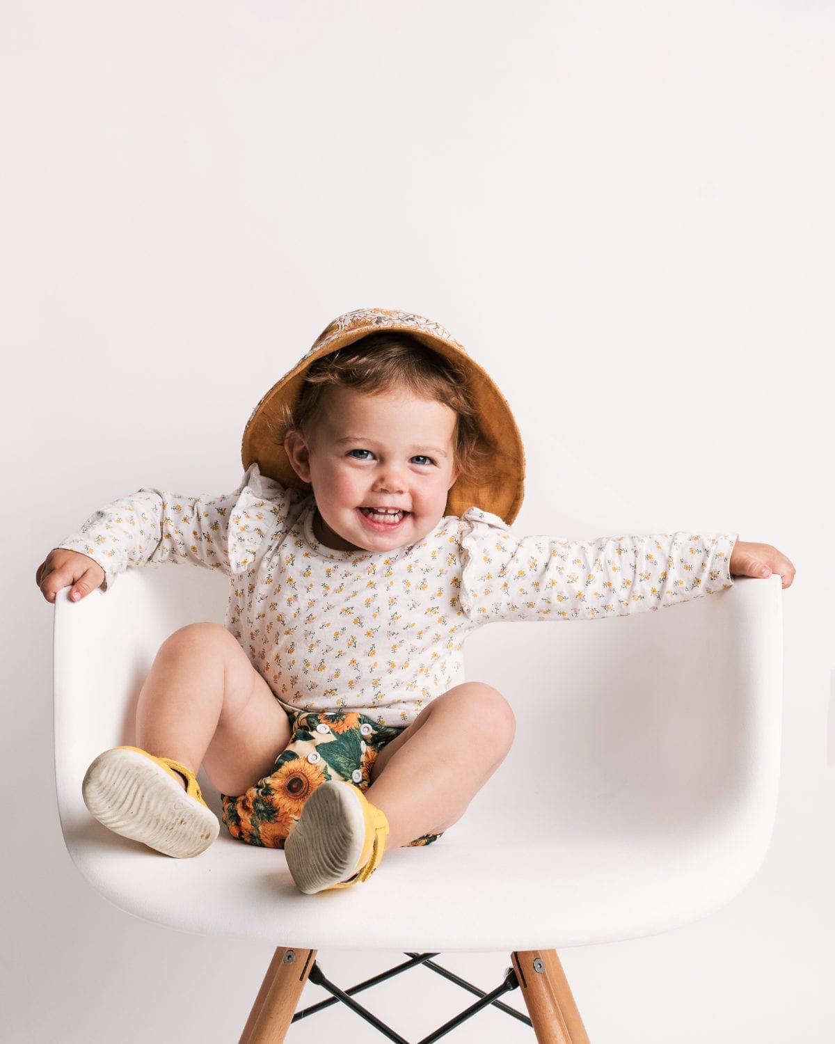 A laughing toddler in a sun hat and sunflower shorts perches on a white designer chair.