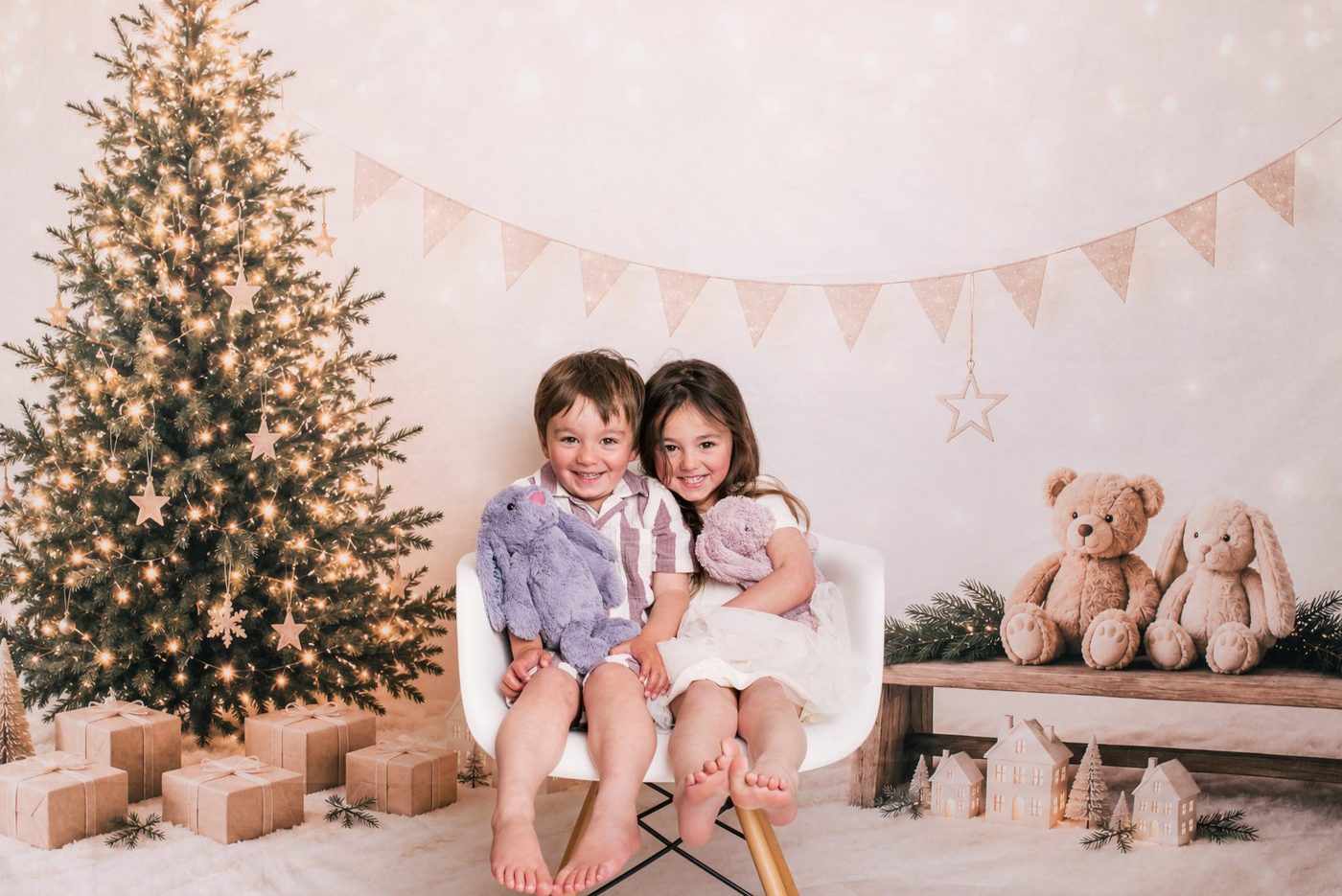 A brother and sister smiling at the camera together on the white chair at this year's Little Portrait Club Christmas session