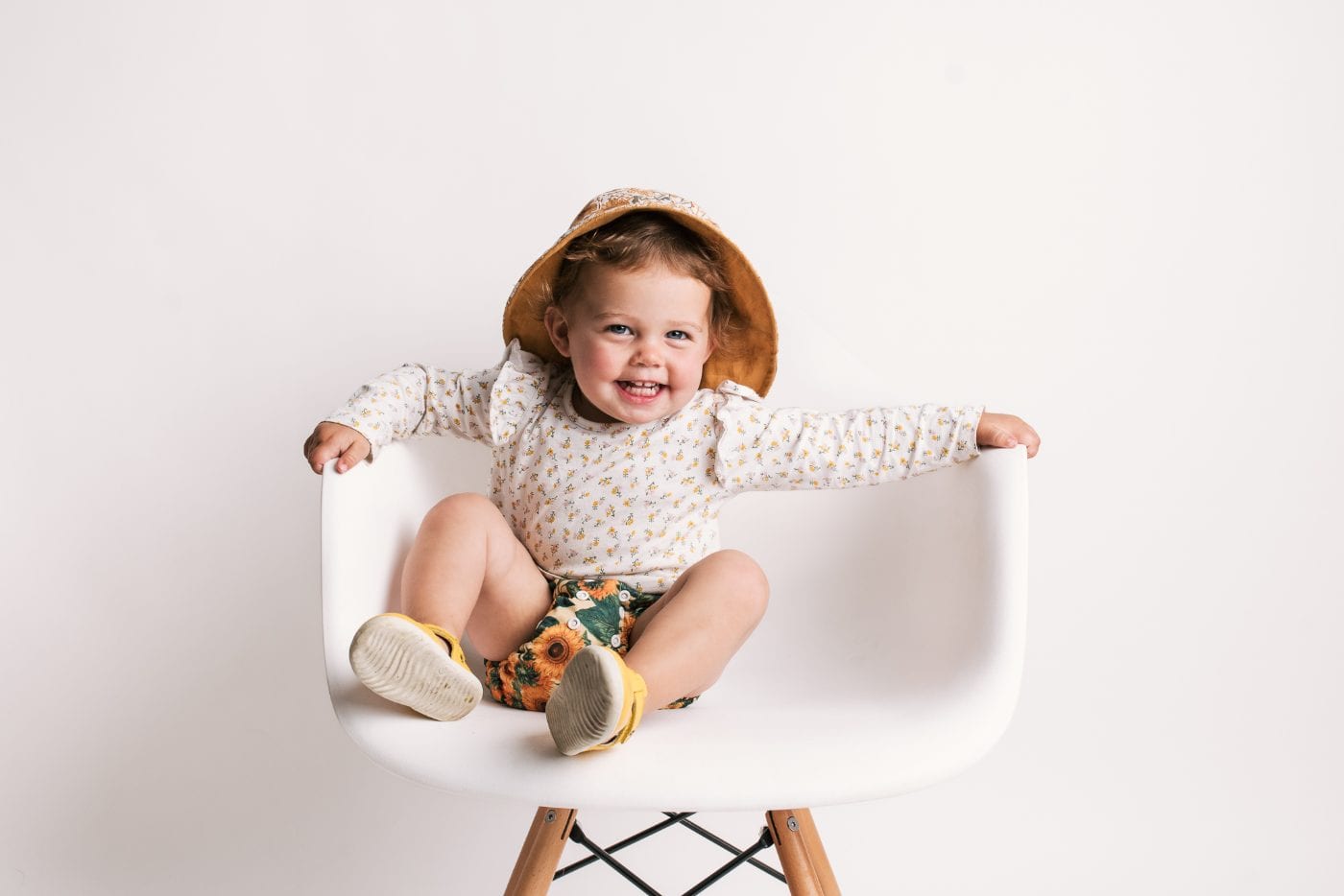 A laughing toddler in a sun hat and sunflower shorts, arms outstretched on either side of the studio chair.