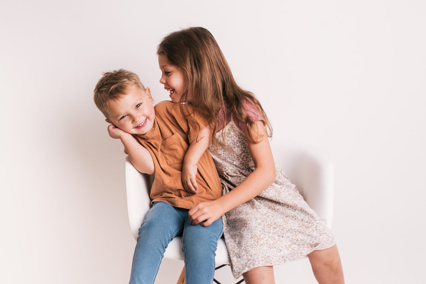 A big sister leaning into her little brother on the studio chair, the two of them laughing together during a Little Portrait Club session.
