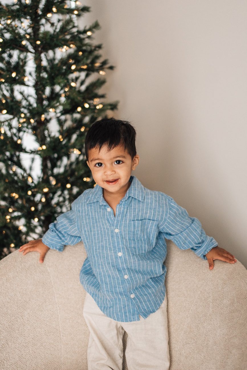 Toddler in a blue linen shirt smiling over the back of a cream sofa, Christmas tree lights behind him