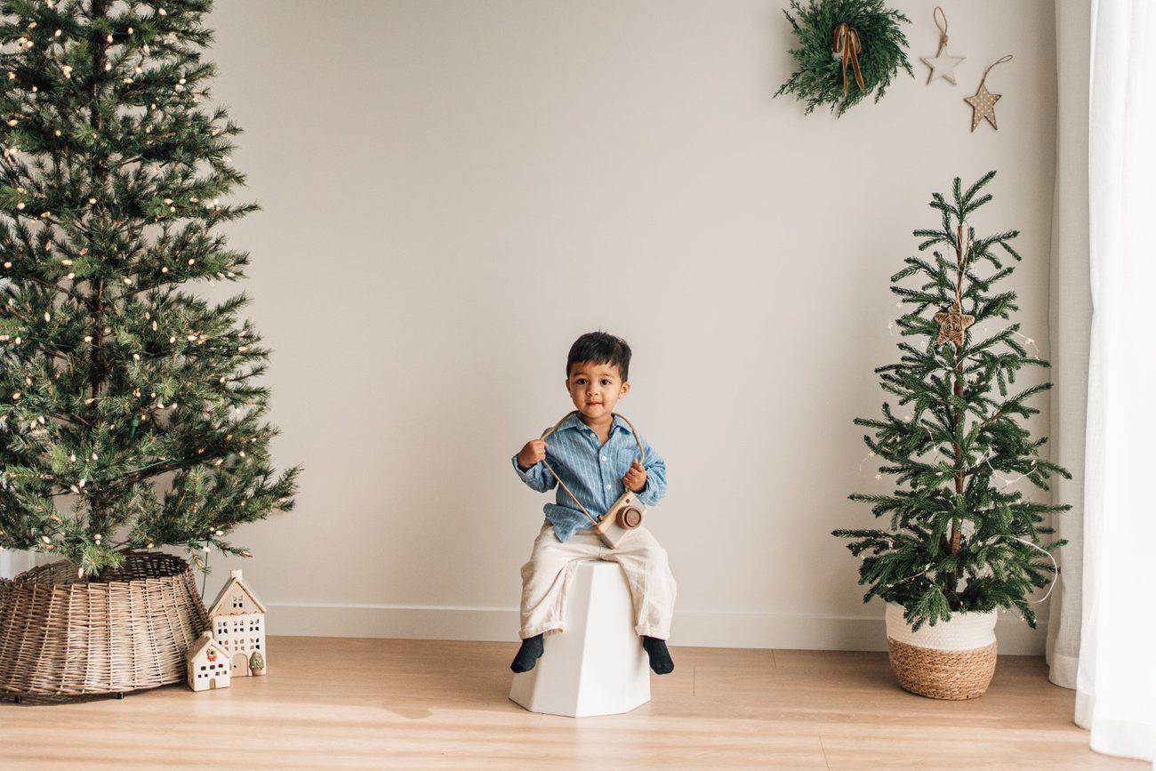 Toddler holding a wooden toy camera, sitting on a white stool between two Christmas trees