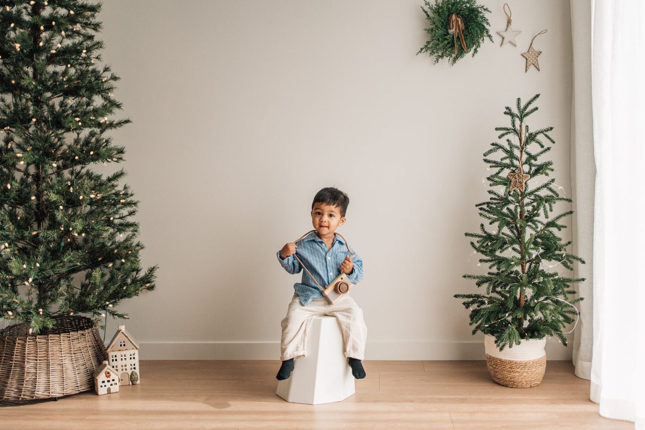 Toddler smiling with a wooden toy camera on a white stool beside the Christmas trees