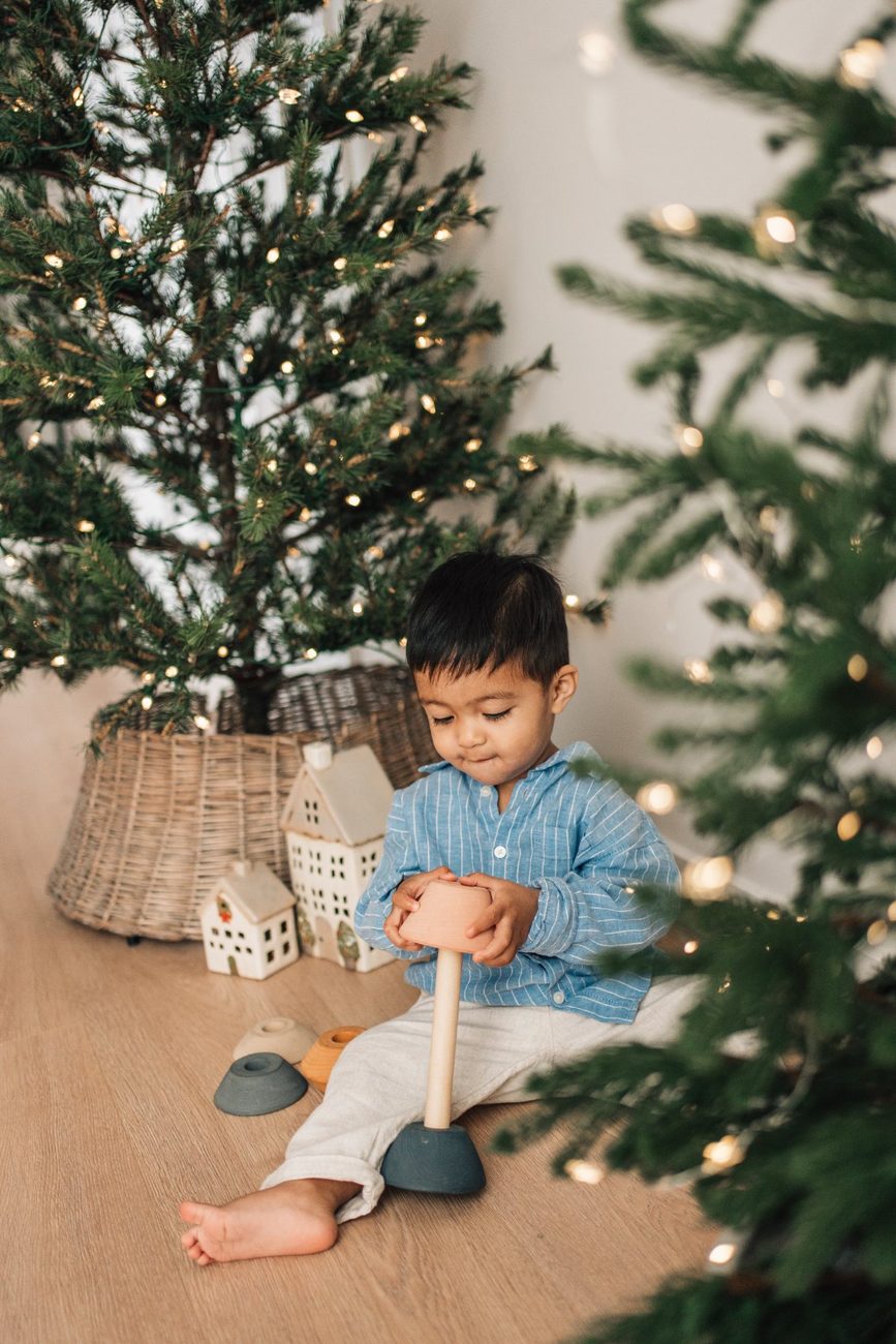 Toddler playing with a wooden stacking toy between two Christmas trees glowing with fairy lights