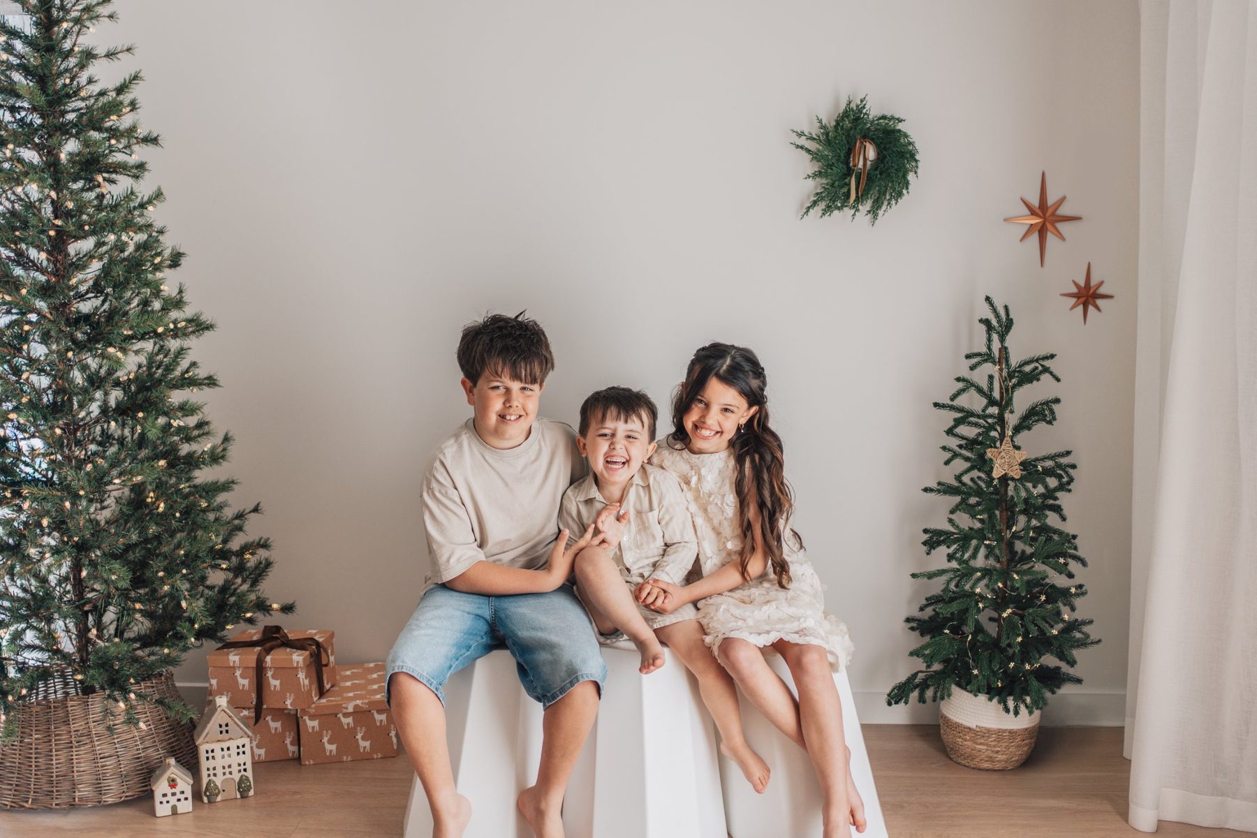 Three siblings laughing together on a white stool between Christmas trees at The Little White Studio