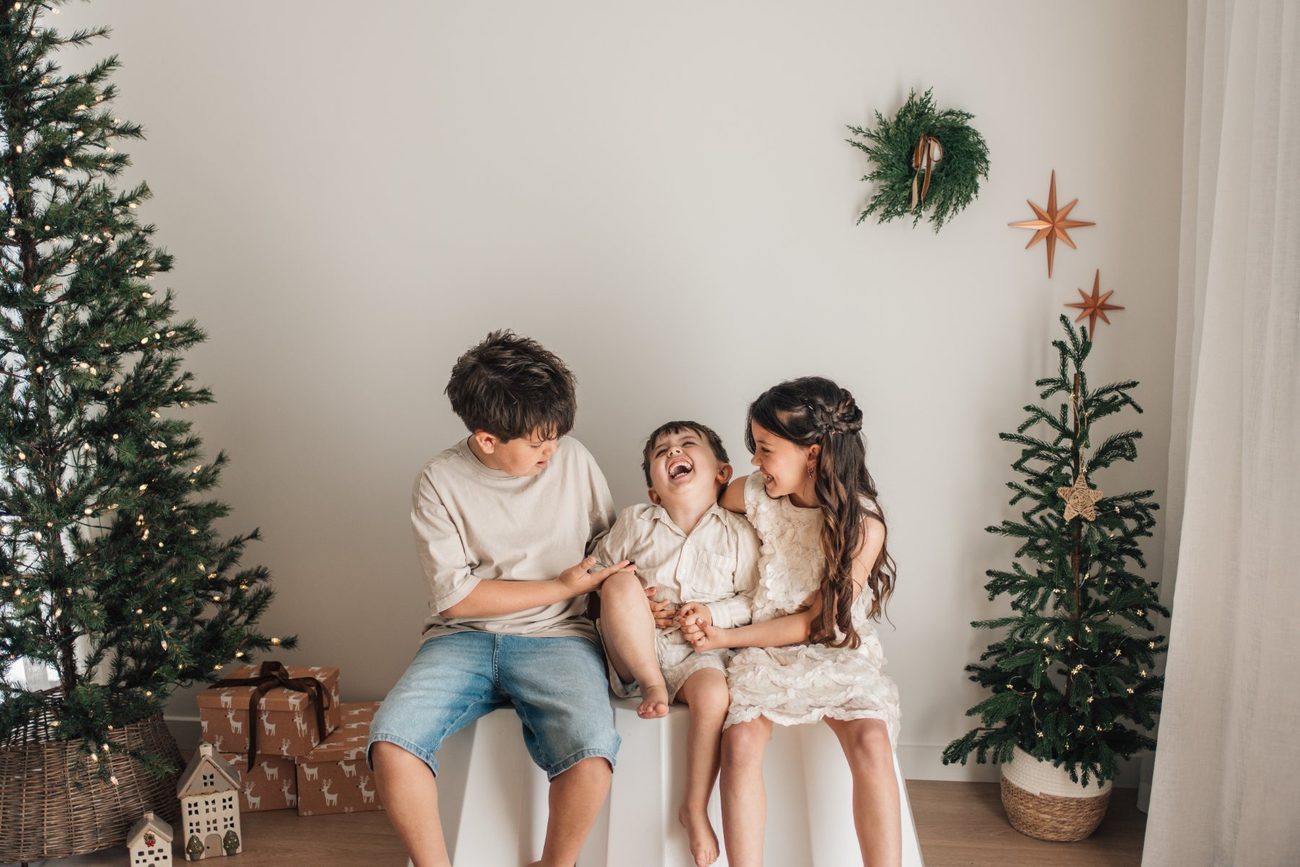 Three siblings laughing together on a white stool between the Christmas trees